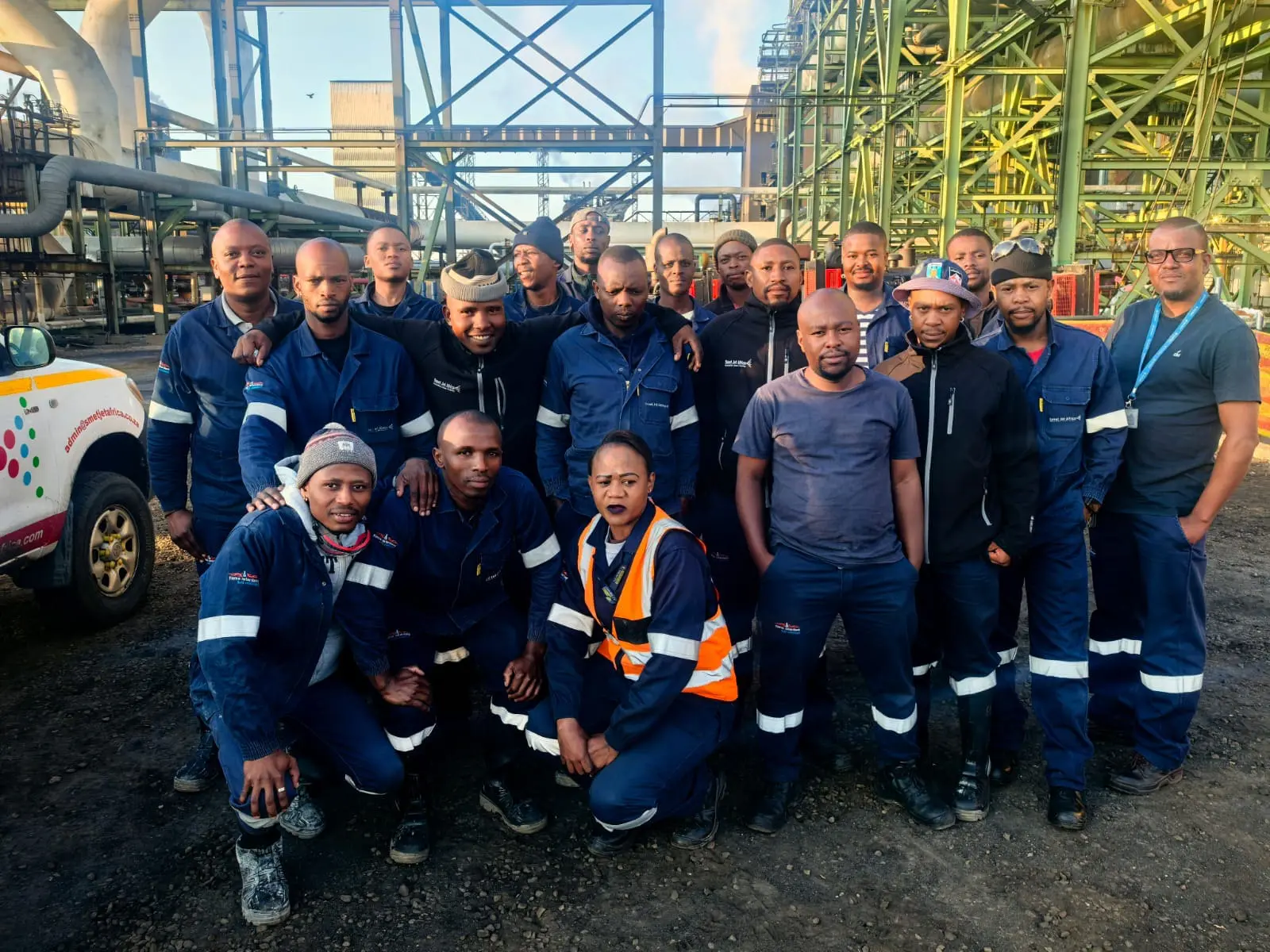 Group of industrial workers in blue uniforms posing outdoors at an industrial facility.