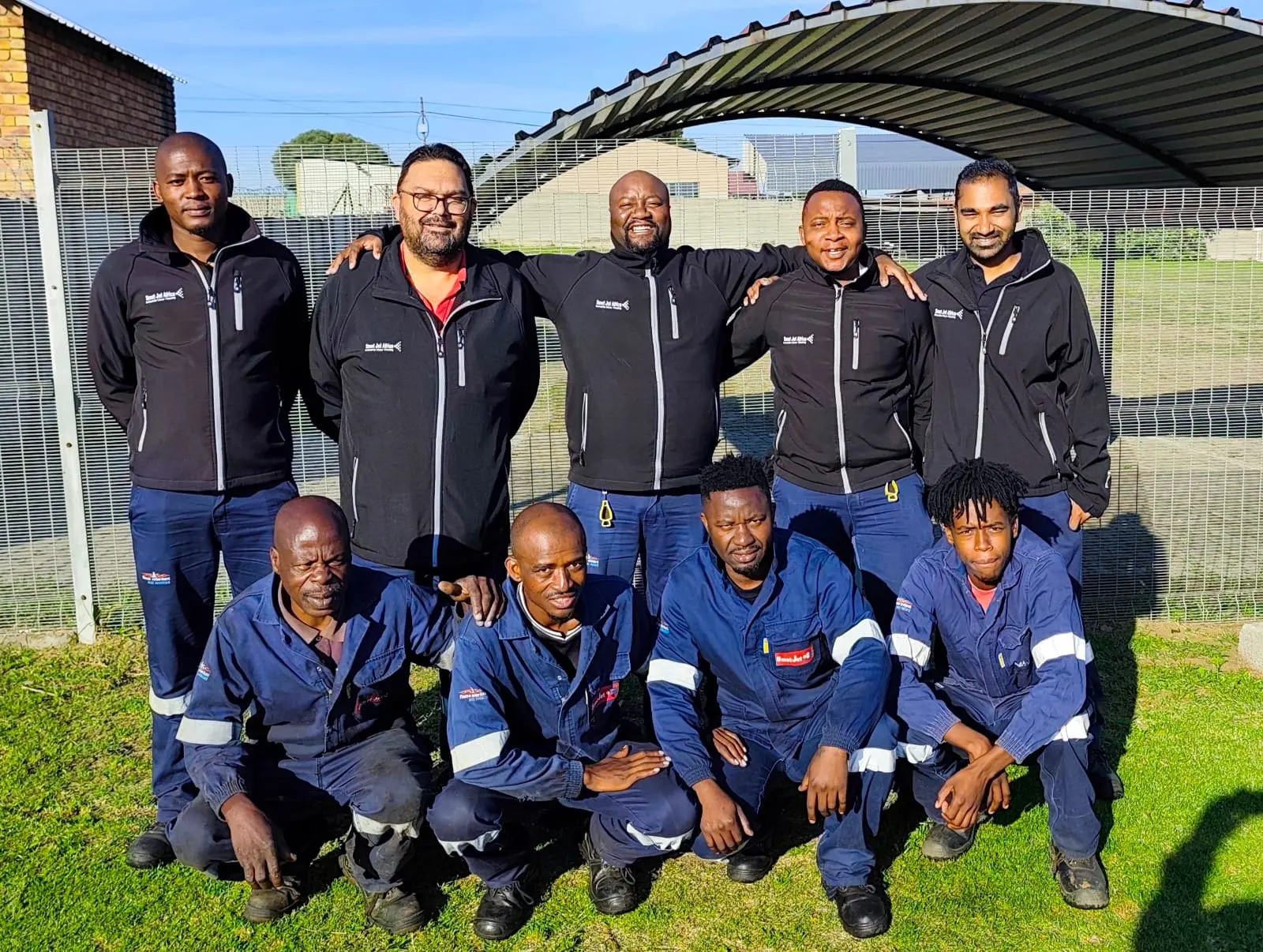 Group of nine men in work uniforms posing outside on grass near fence under blue sky.