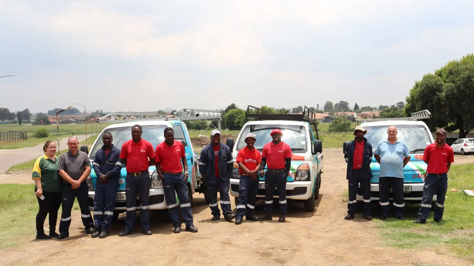 Group of eleven workers standing in front of three company vans on a dirt road outdoors.