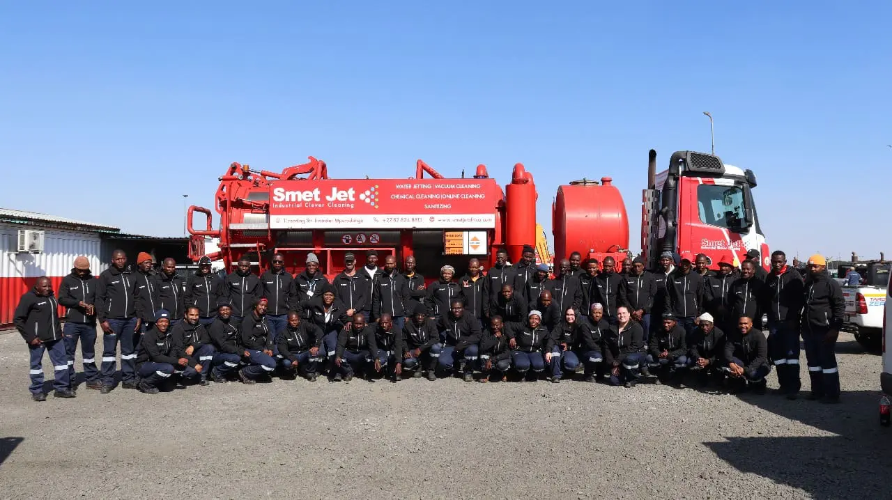 Large group of workers in black jackets posing in front of a red Smet Jet industrial cleaning truck.