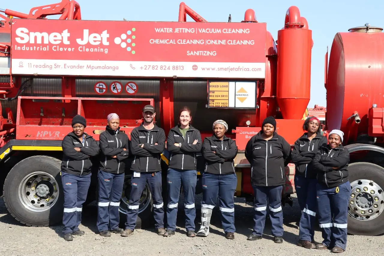 Eight workers in uniform standing with arms crossed in front of a red Smet Jet industrial cleaning truck.