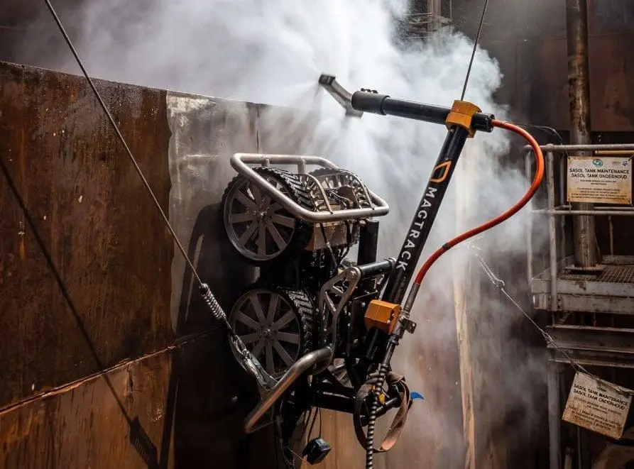 Robot with tracks inspecting a rusty metal wall emitting white smoke or steam indoors.