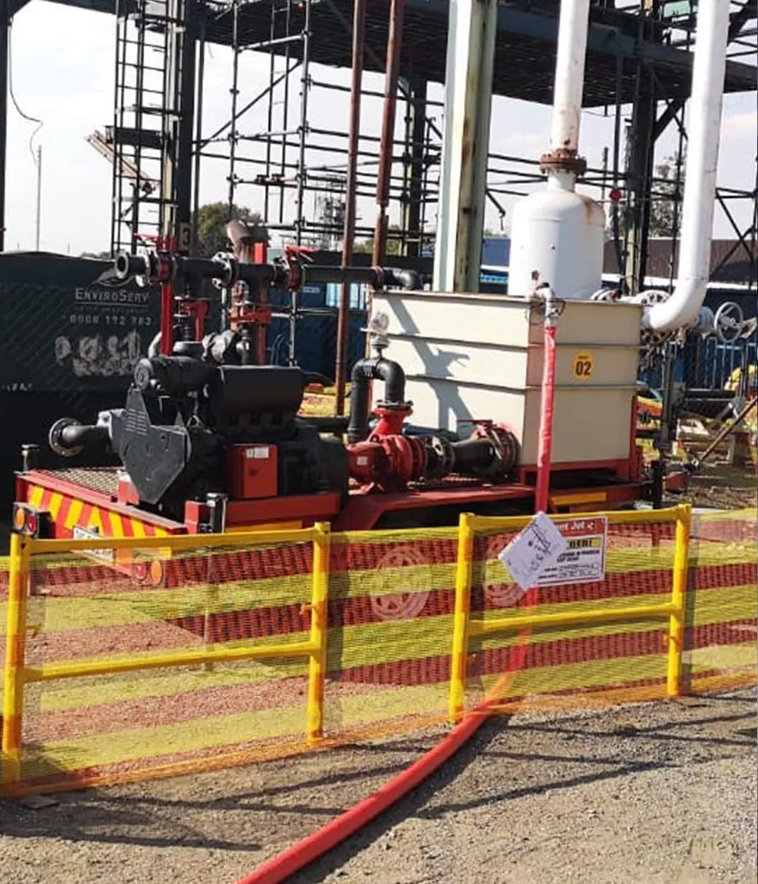 Industrial pump and piping system surrounded by yellow and red safety fencing on gravel ground.