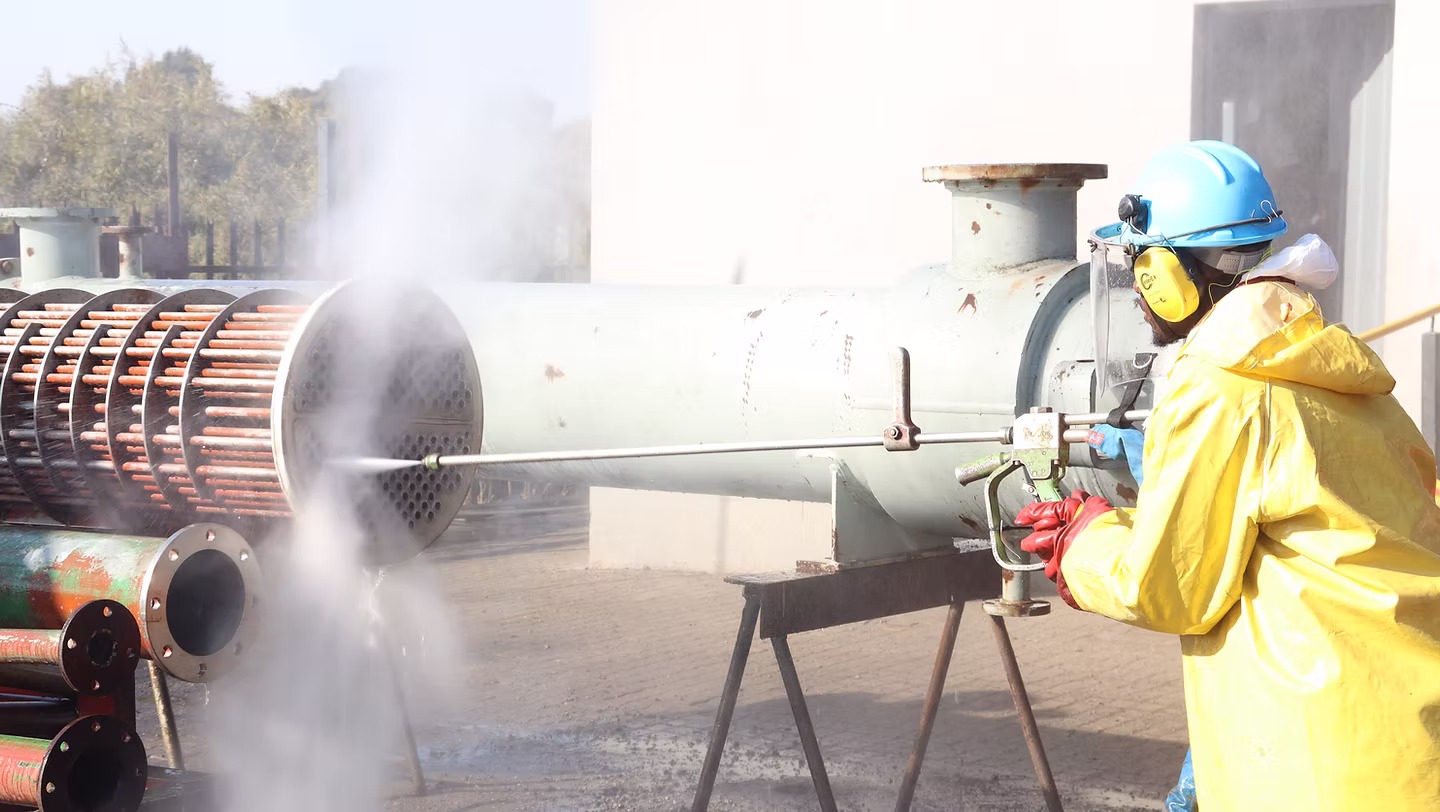 Worker in safety gear using high-pressure water to clean industrial heat exchanger tubes outdoors.