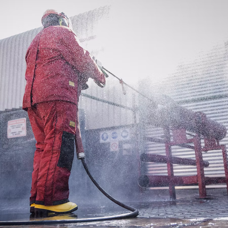 Person in red protective gear and yellow boots spraying water with a hose near industrial pipes.