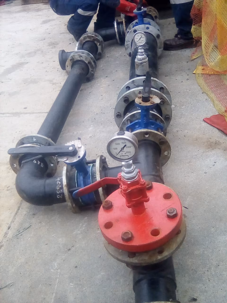 Industrial pipes with pressure gauges and valves on a concrete floor, workers partly visible.
