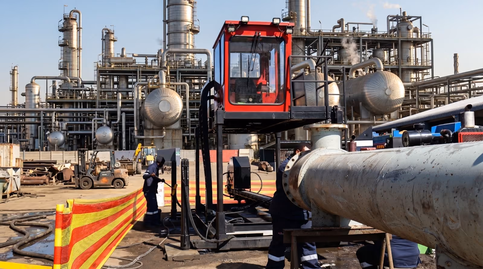 Workers operating industrial machinery with large pipes inside a factory complex with metal structures and tanks.