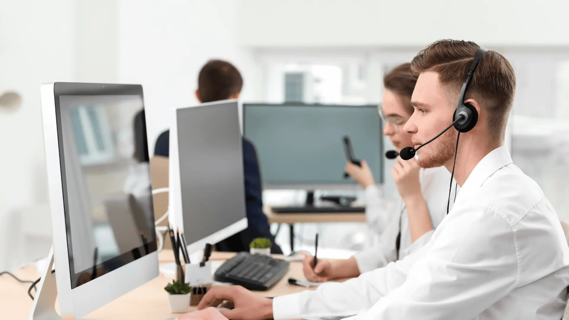 Jeune homme portant un casque avec microphone utilise un ordinateur dans un bureau, d'autres personnes travaillent en arrière-plan avec des casques.