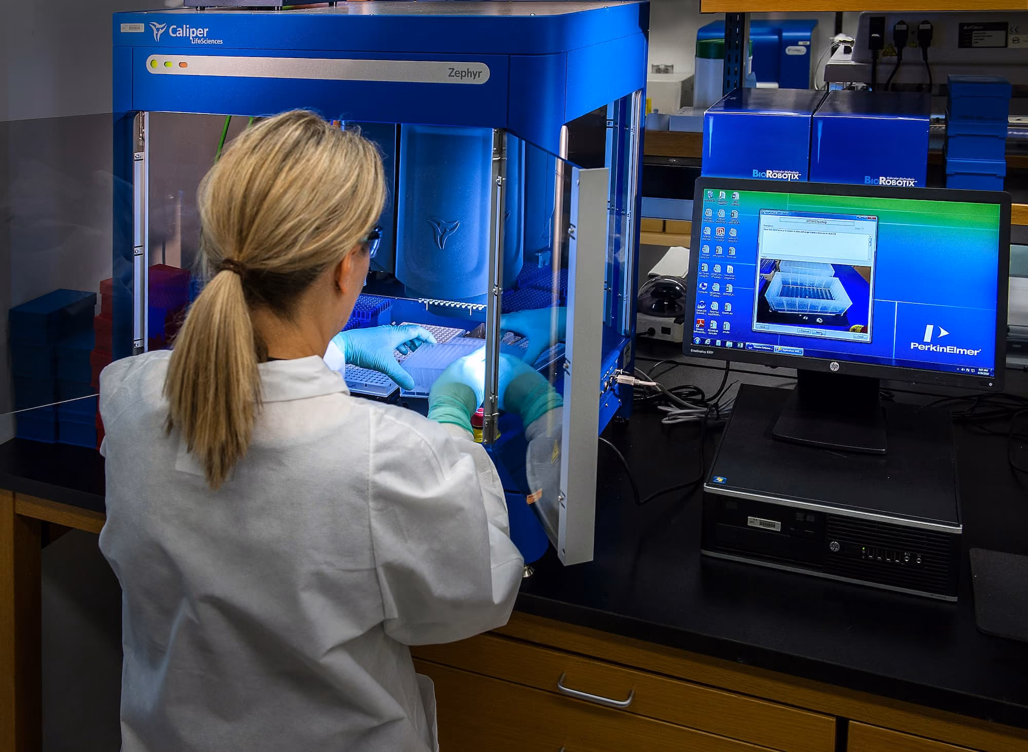 Une scientifique portant une blouse blanche et des gants bleus manipule des échantillons dans un appareil bleu Caliper Zephyr en laboratoire, avec un ordinateur affichant le logiciel PerkinElmer à côté.