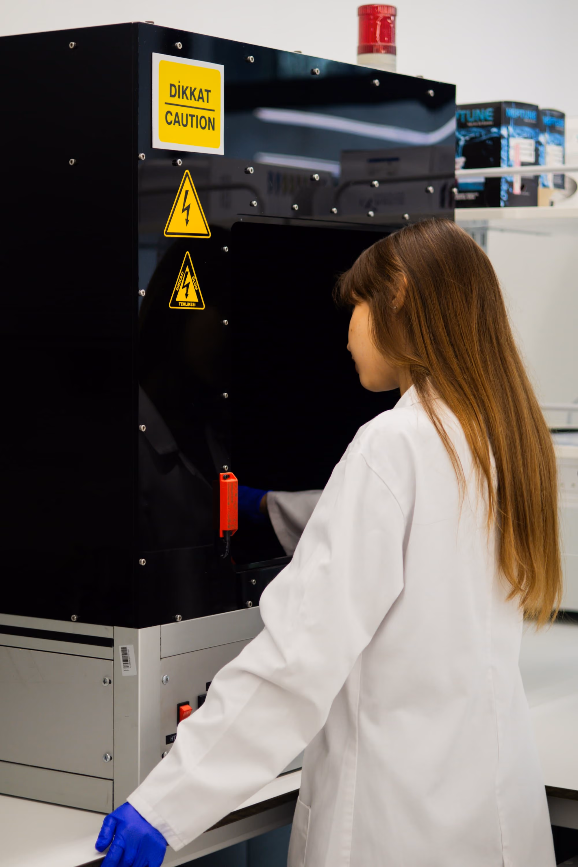 Une femme en blouse blanche et gants bleus utilise une machine noire dans un environnement de laboratoire, avec des panneaux de mise en garde jaune indiquant un risque électrique.