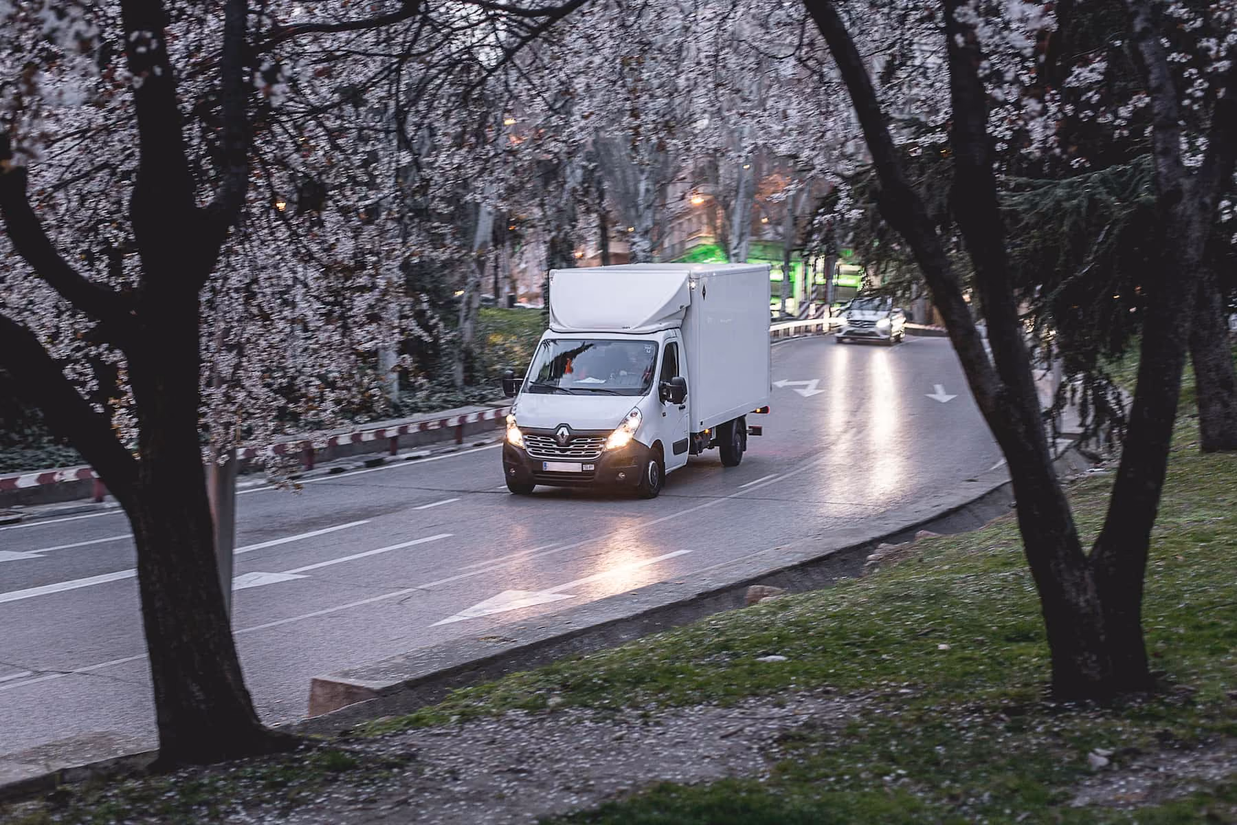 Un camion blanc avec phares allumés roulant sur une route bordée d'arbres en fleurs au crépuscule.