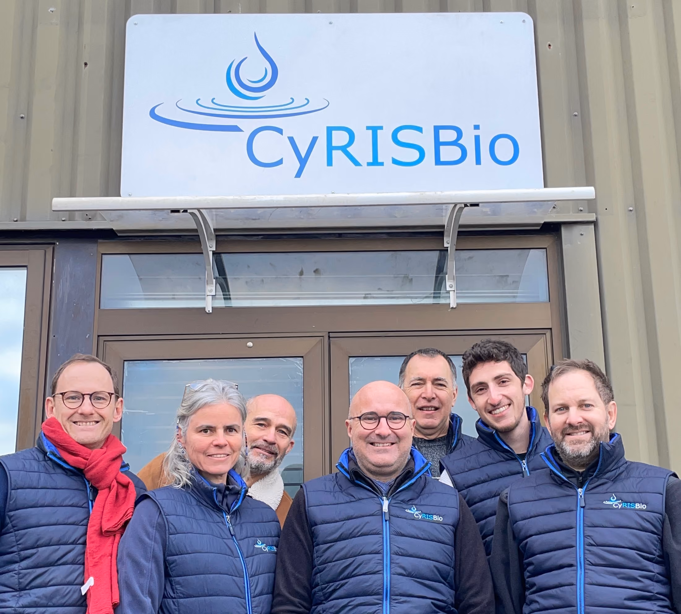 Un groupe de sept personnes portant des vestes bleu marine avec le logo CyRISBio, posant devant la porte d'entrée d'un bâtiment sous une enseigne avec le même logo bleu et turquoise.