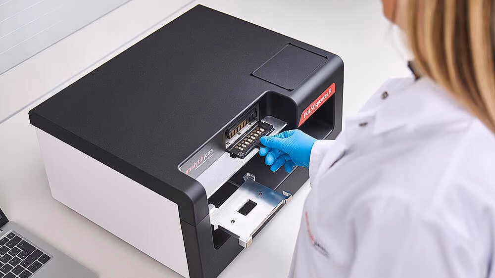 Une personne en blouse blanche et gants bleus insère une platine dans un appareil de laboratoire noir et blanc nommé PULSEspencer R.