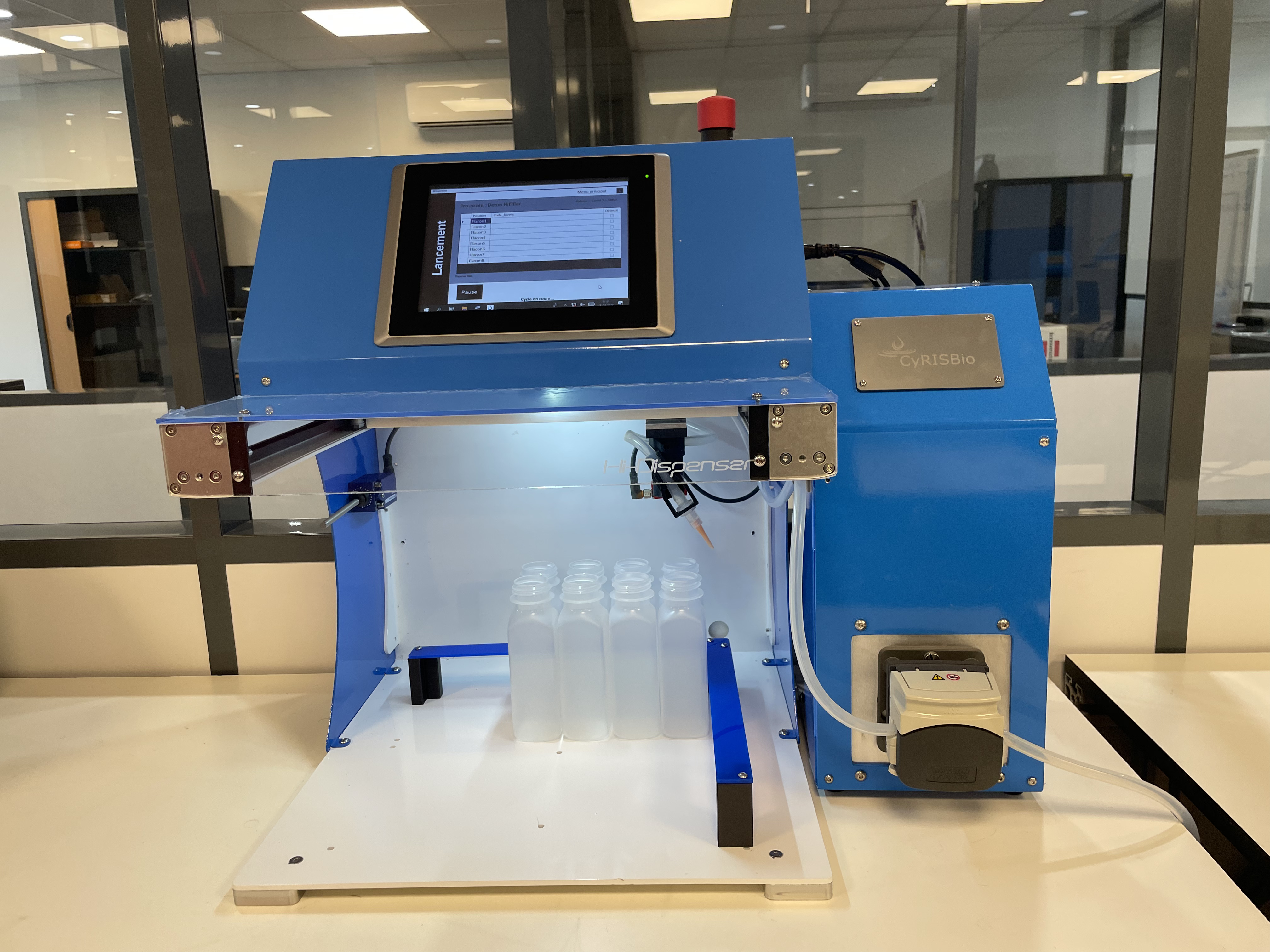 Distributeur automatique de liquide bleu et blanc sur un bureau, équipé d'un écran tactile affichant un menu de lancement, avec plusieurs bouteilles transparentes placées sous la buse de distribution.