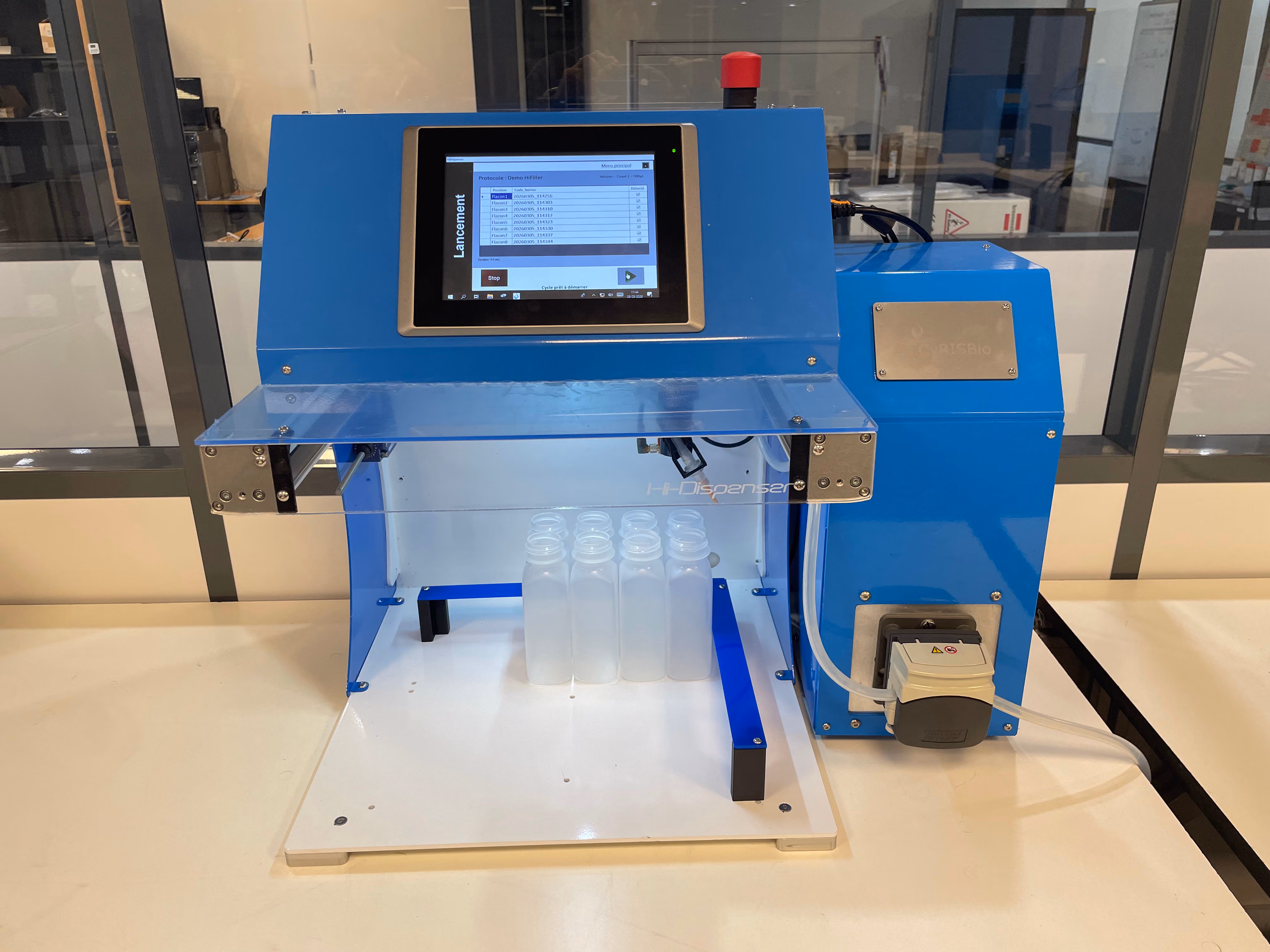 Machine bleue de laboratoire avec écran tactile affichant une interface utilisateur pour le lancement d’un processus, plusieurs bouteilles en plastique transparent placées sous un compartiment protégé par une plaque transparente, sur une table blanche dans une pièce équipée de grandes fenêtres.