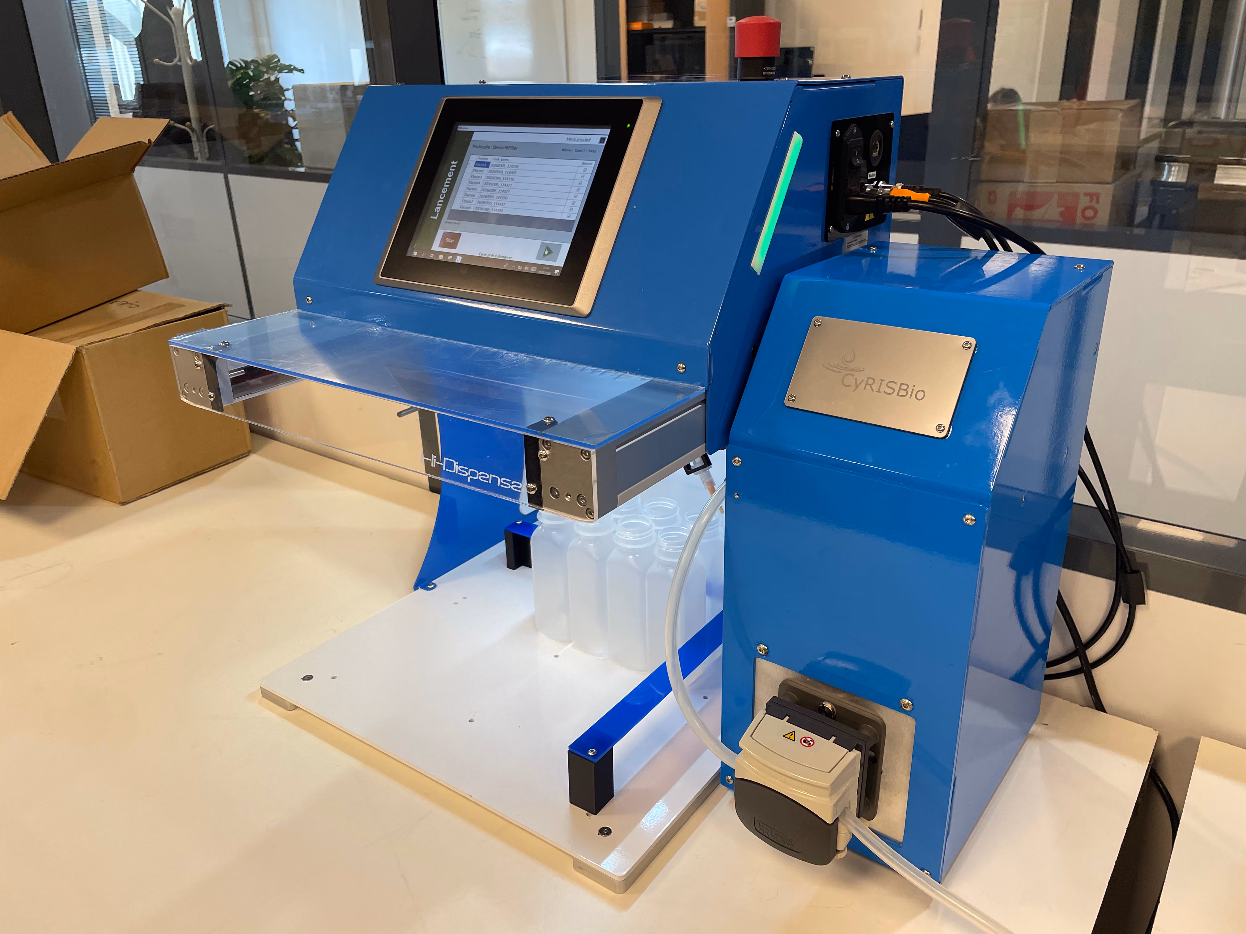Appareil de laboratoire bleu avec écran tactile affichant un logiciel pour gérer un protocole, étiqueté Hi-Dispense et CyRISBio, avec plusieurs petites bouteilles transparentes alignées en dessous, posé sur une surface blanche dans un environnement de laboratoire.