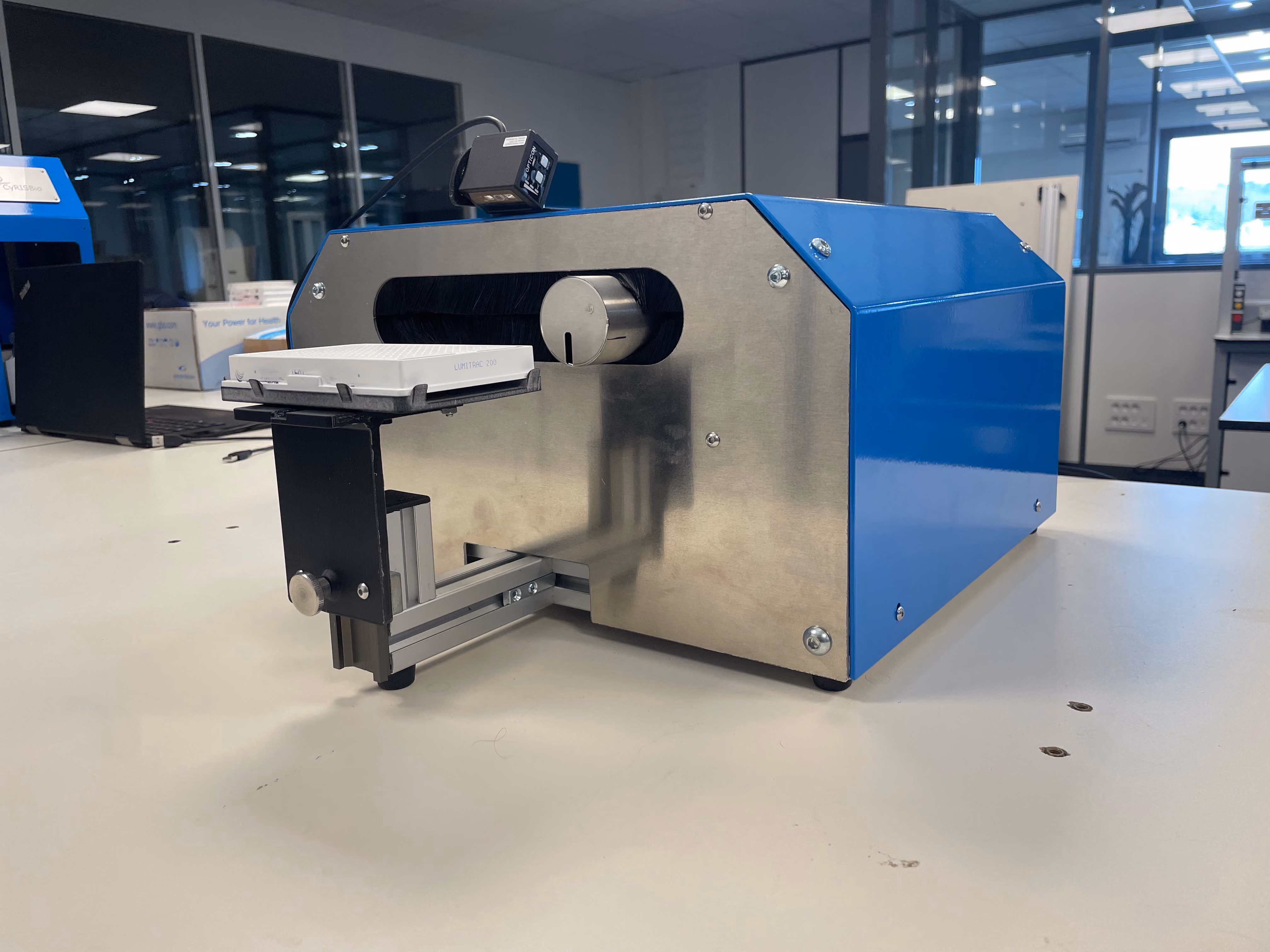 Machine de laboratoire métallique et bleue avec un plateau coulissant noir tenant une plaque blanche étiquetée LUMITRAC 200, sur une table blanche dans un laboratoire moderne avec des fenêtres et un ordinateur portable en arrière-plan.