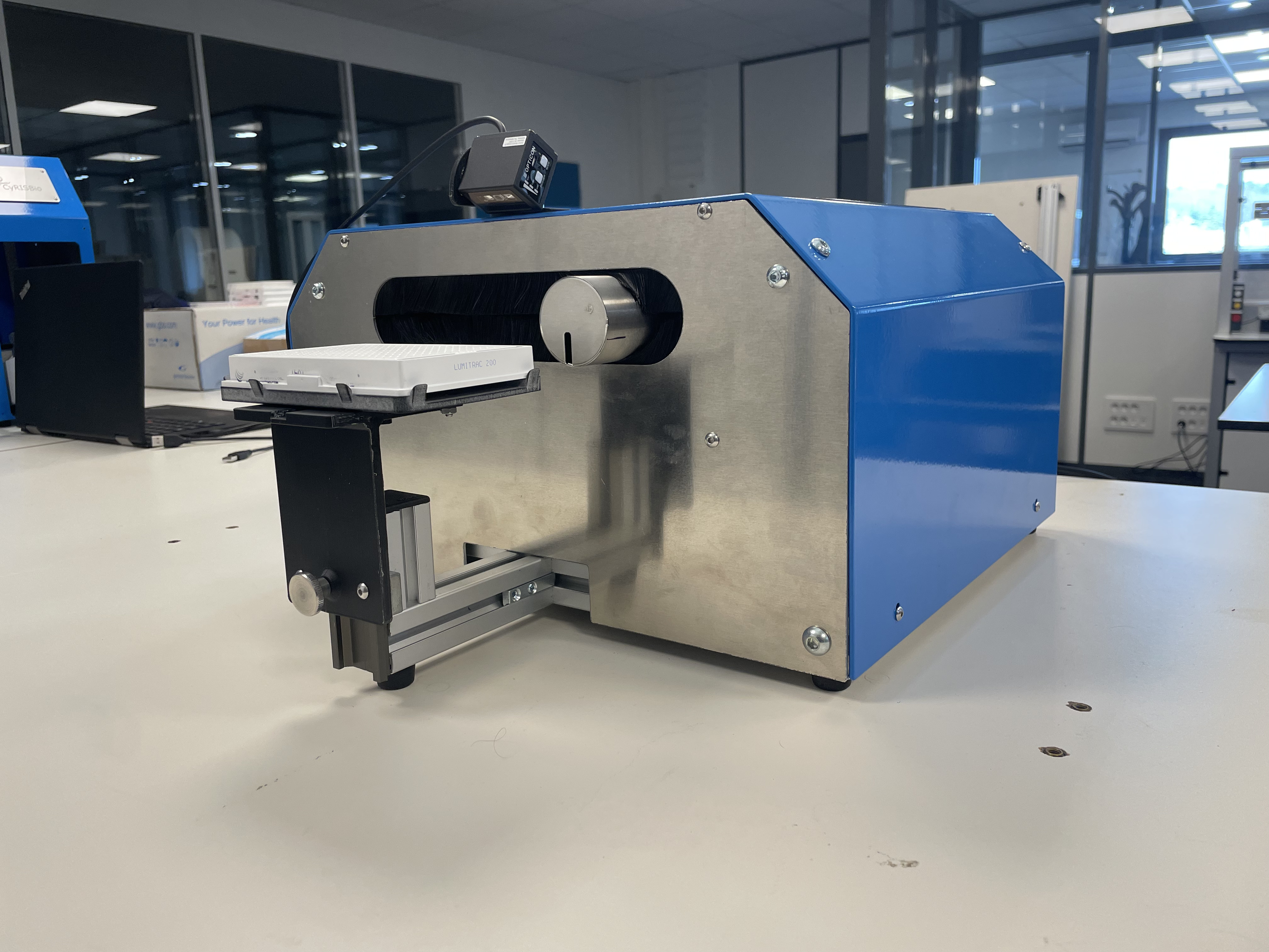 Machine de laboratoire métallique et bleue avec un plateau coulissant noir tenant une plaque blanche étiquetée LUMITRAC 200, sur une table blanche dans un laboratoire moderne avec des fenêtres et un ordinateur portable en arrière-plan.