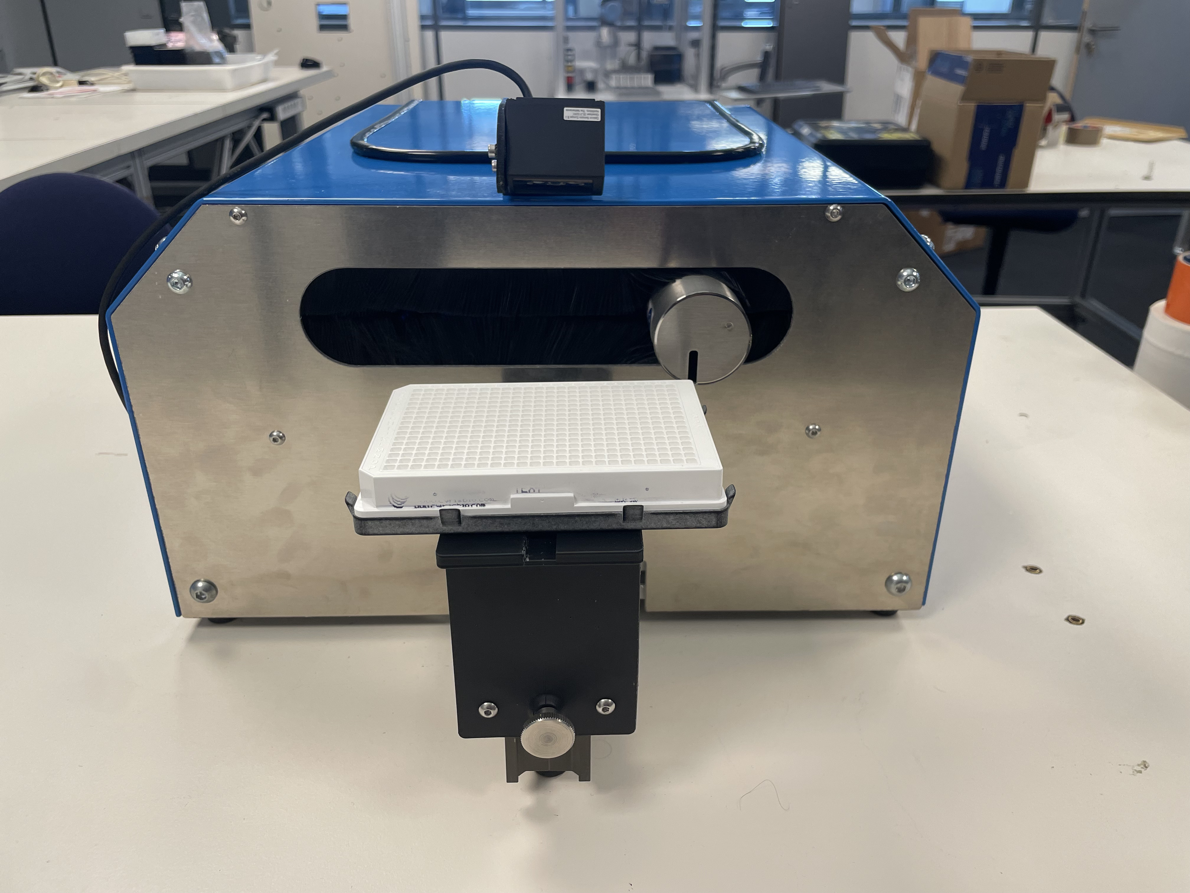 Appareil de laboratoire bleu et argent avec un plateau blanc quadrillé fixé à un support noir, installé sur une table blanche dans une pièce avec des bureaux et des boîtes en arrière-plan.