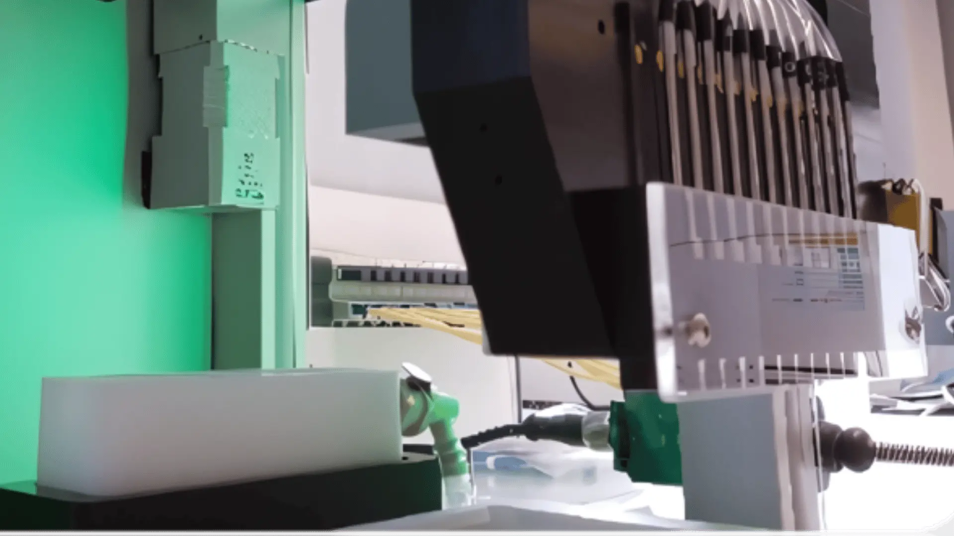 Distributeur liquide avec plusieurs têtes interchangeables en métal et plastique, équipé de plusieurs tubes jaunes connectés à un système de distribution, sur un fond vert et un bureau blanc.