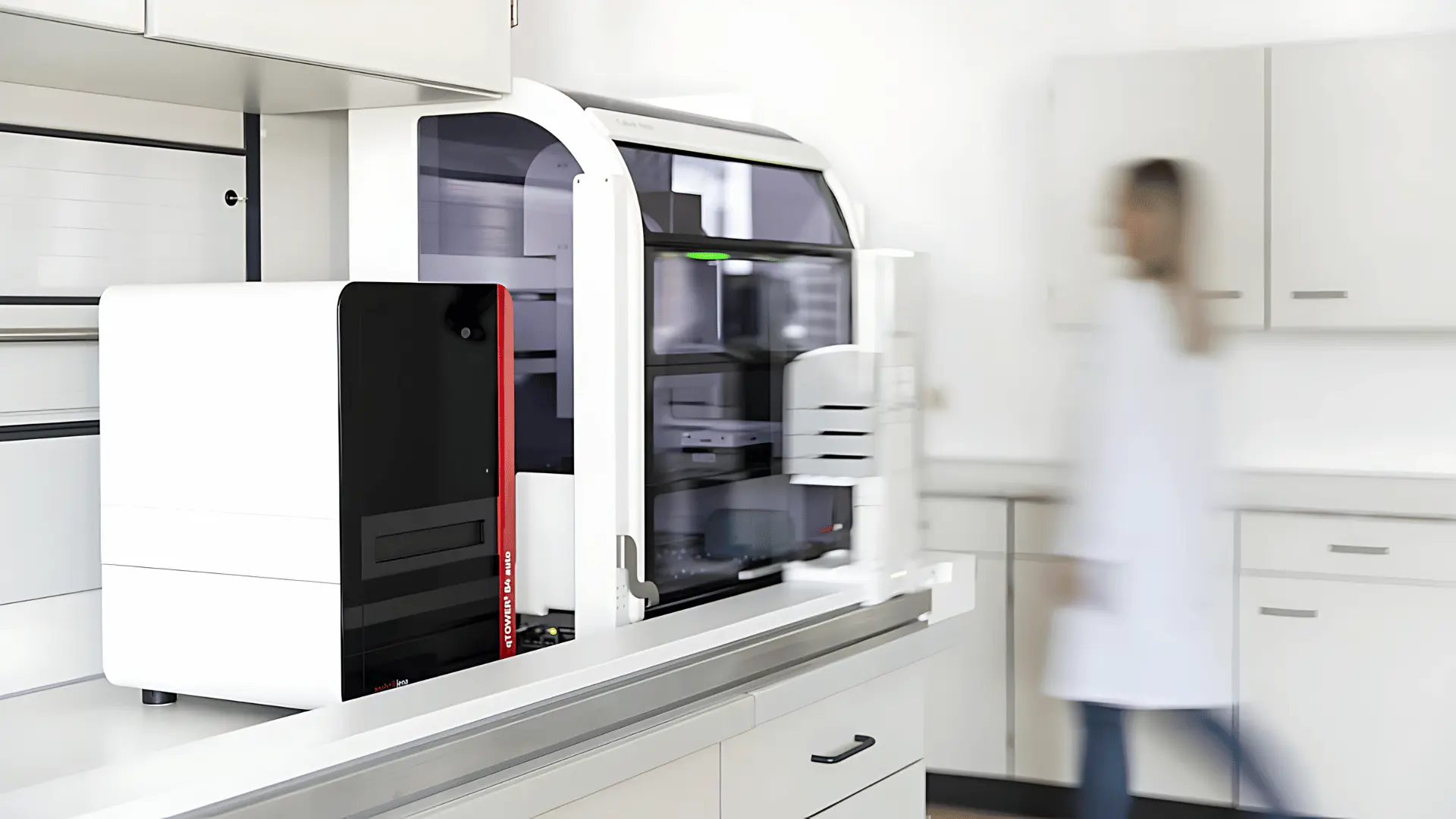 Appareil médical automatisé blanc et noir avec une porte vitrée noircie dans un laboratoire propre, une personne portant une blouse blanche est floue en arrière-plan.