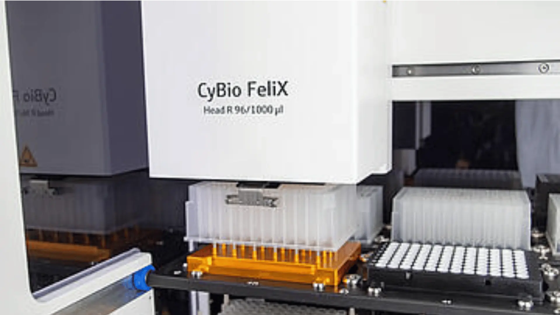 Partie d'un appareil de laboratoire automatisé CyBio FeliX, montrant des plaques de microtubes et une tête de pipette pour manipulation de liquides.