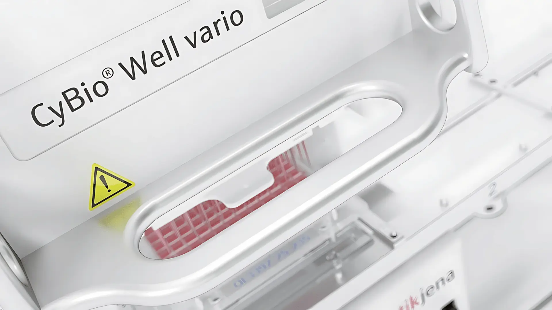 Détail d'un instrument de laboratoire CyBio Well vario avec un avertissement jaune triangulaire et des pointes de pipette rouges visibles à travers une ouverture.