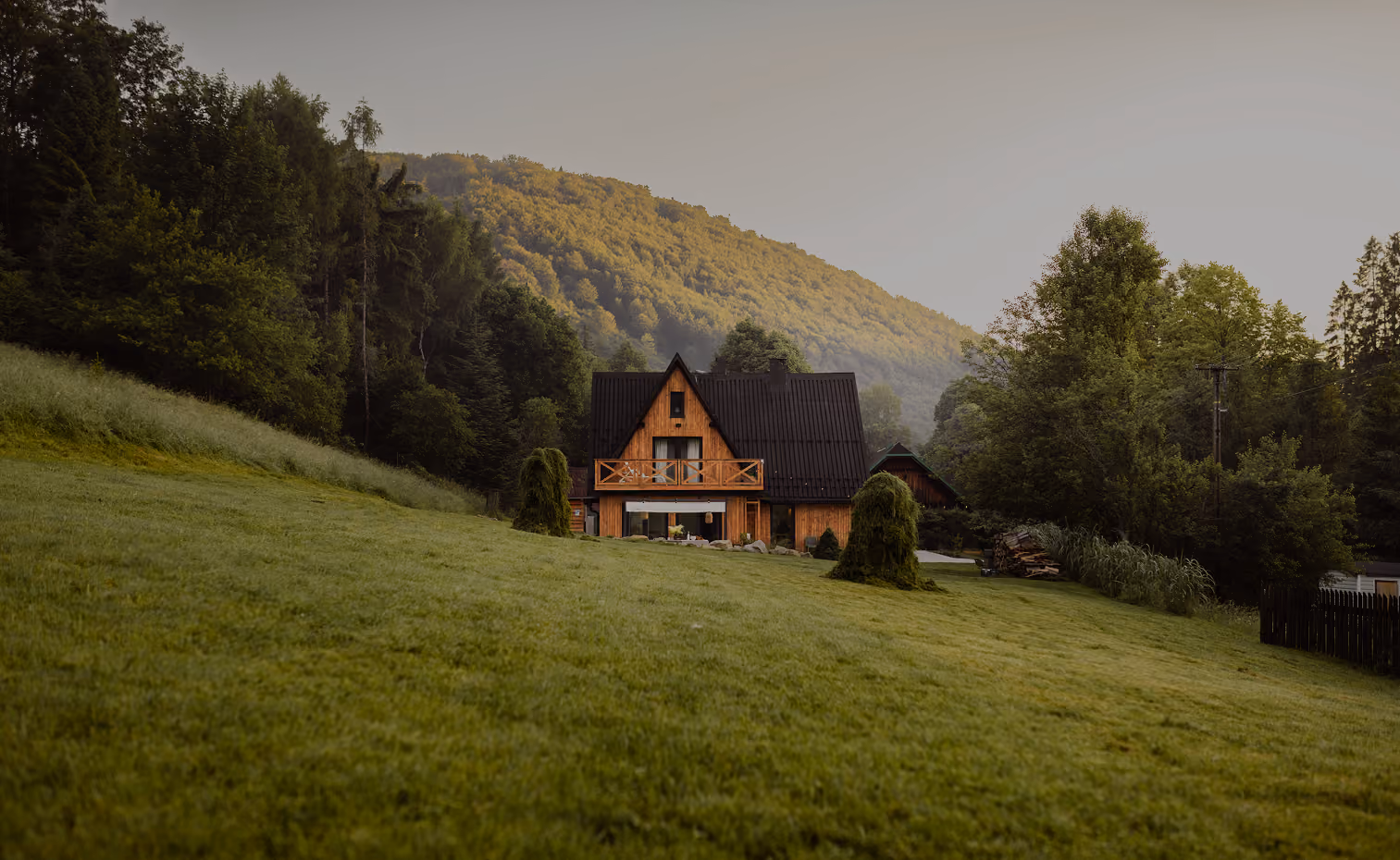 Wood cabin with black roof on grassy hill, surrounded by trees and forested hills in background.