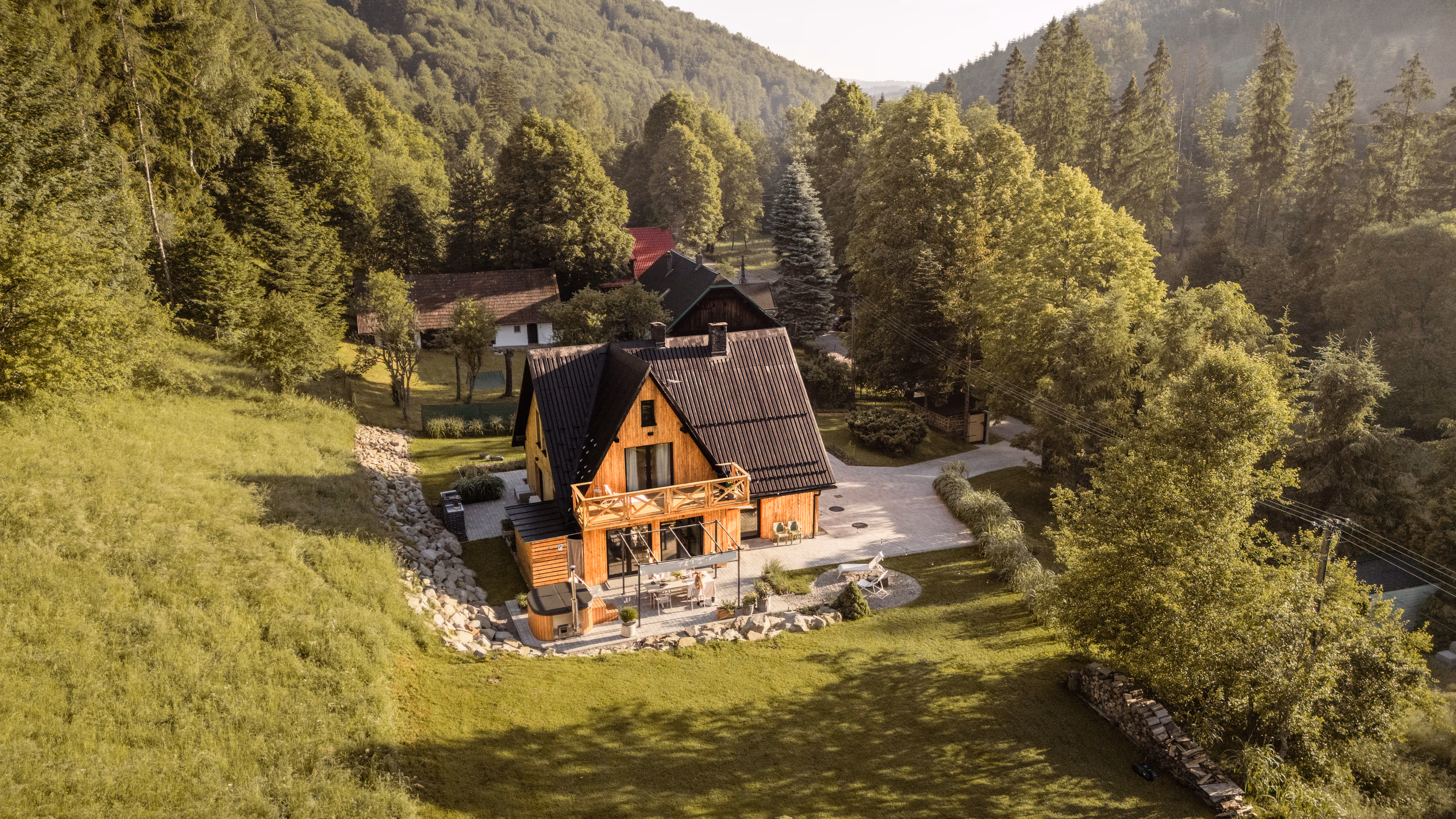Wooden house with a black roof in a green valley surrounded by trees and hills.
