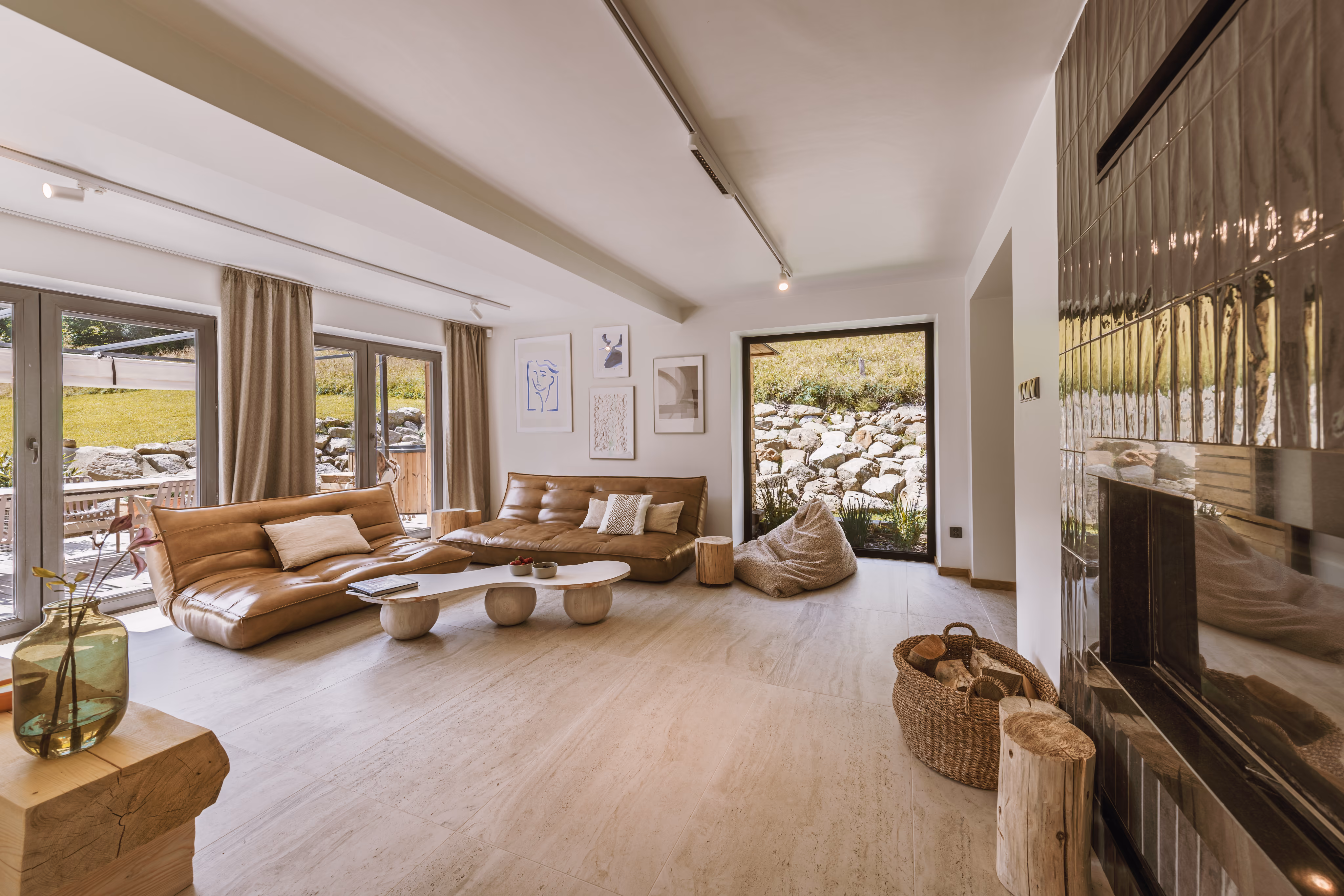 Modern living room with two brown sofas, round coffee tables, large windows, and a basket of firewood.