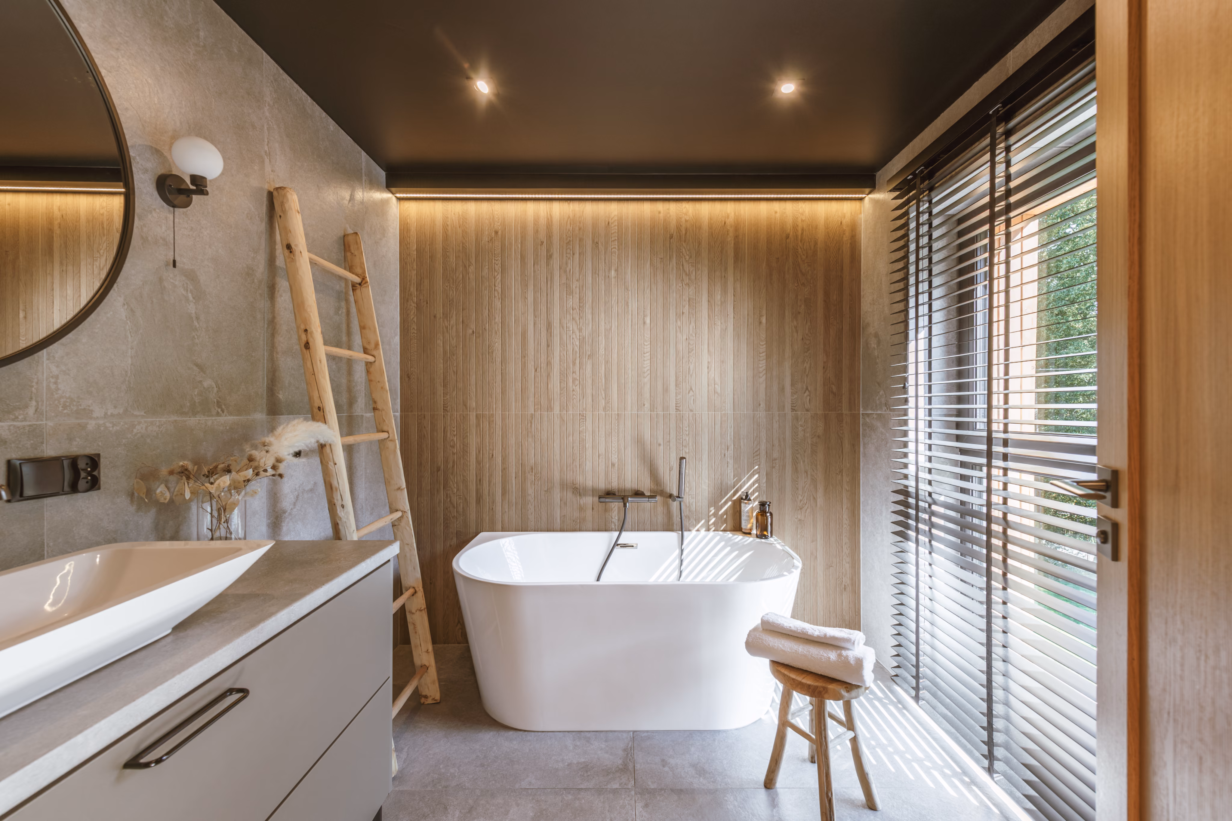Modern bathroom with white freestanding tub, wooden wall, ladder, vanity, and window blinds letting in sunlight.