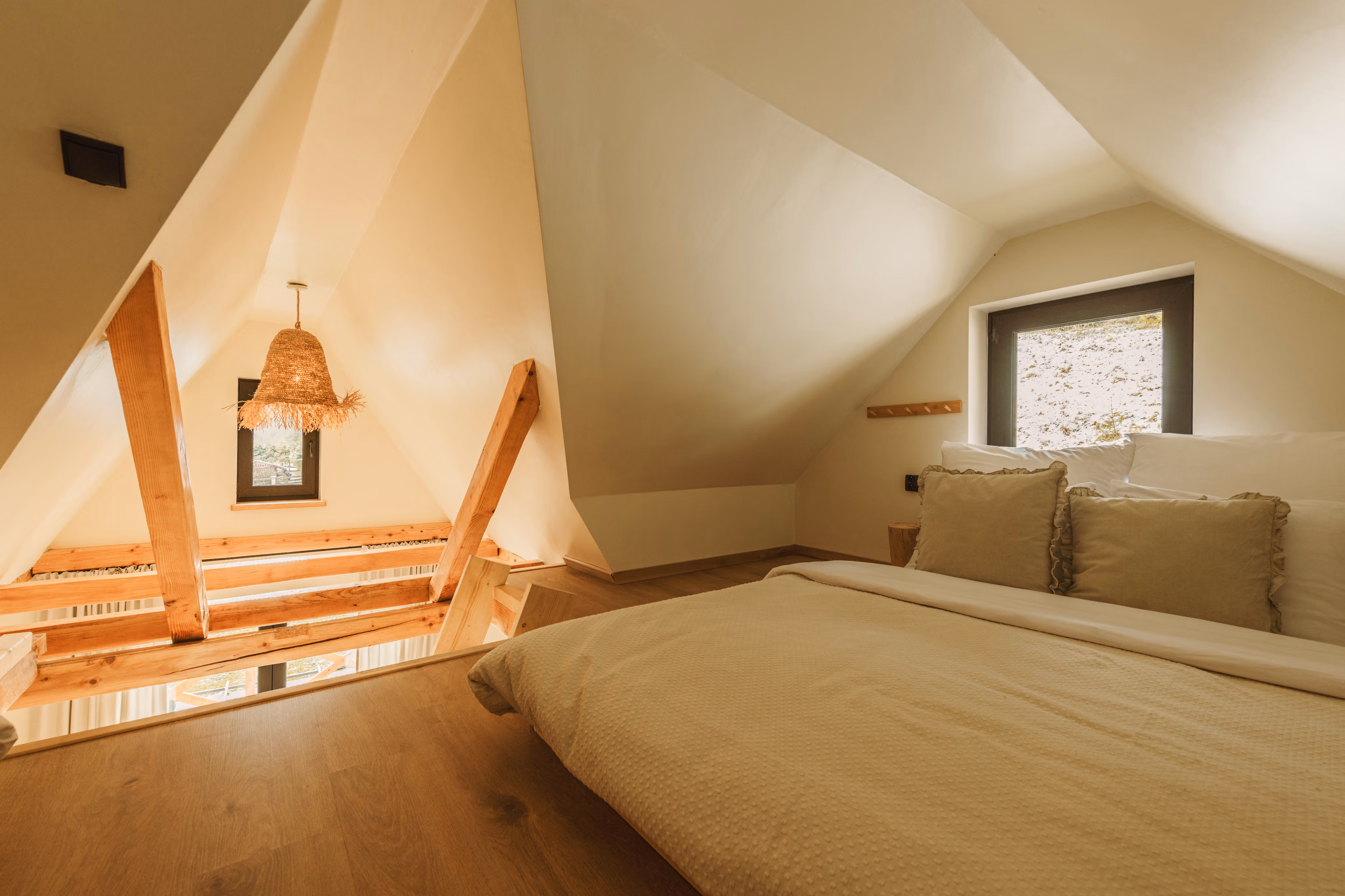 Cozy attic bedroom with beige bedding, ruffled pillows, sloped ceiling, wooden beams, and small windows.