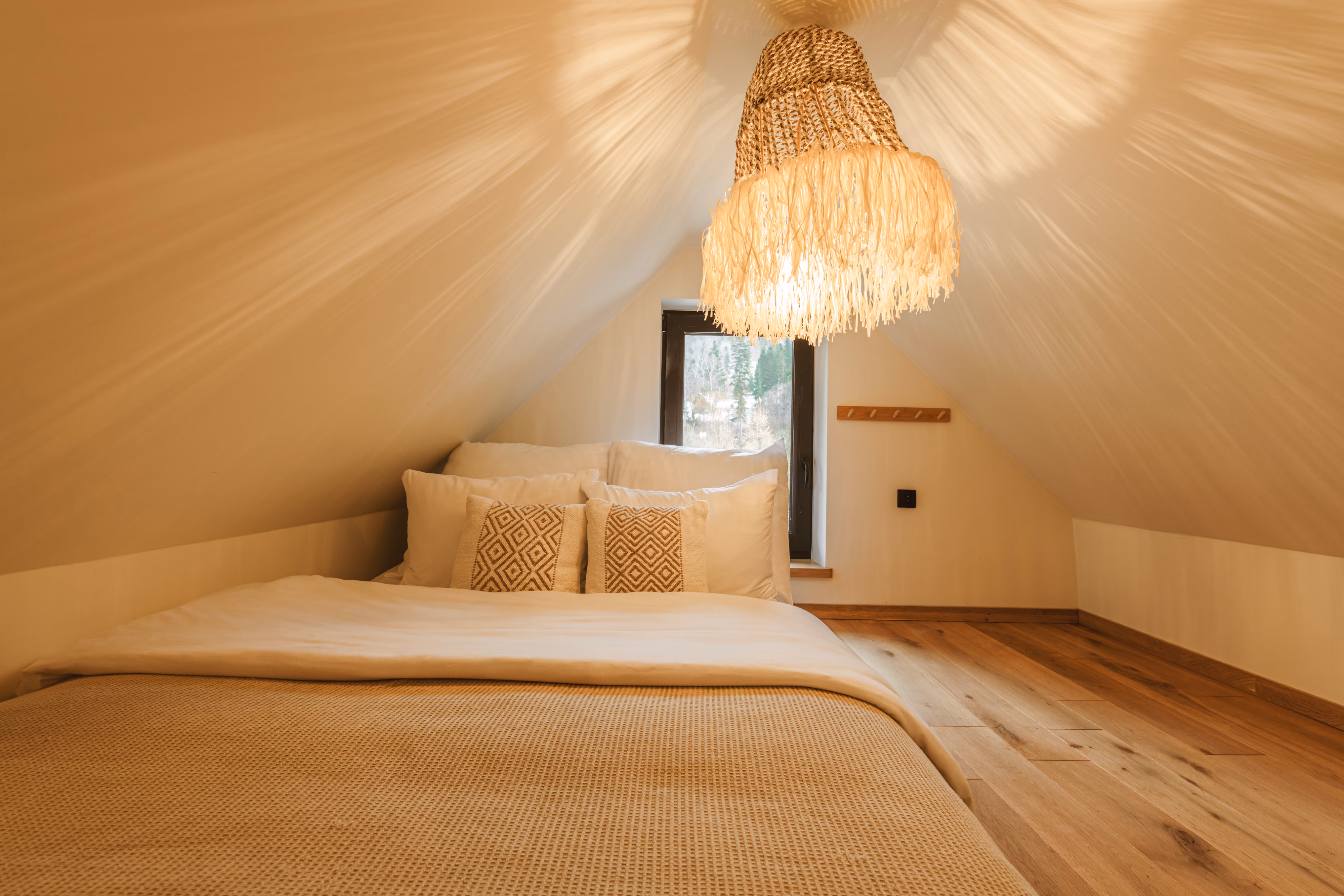 Cozy attic bedroom with white bedding, patterned pillows, wooden floor, and woven hanging light fixture.