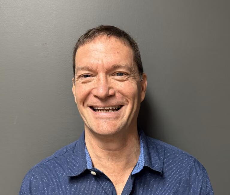 Smiling middle-aged man with short hair wearing a blue patterned button-up shirt against a gray wall.