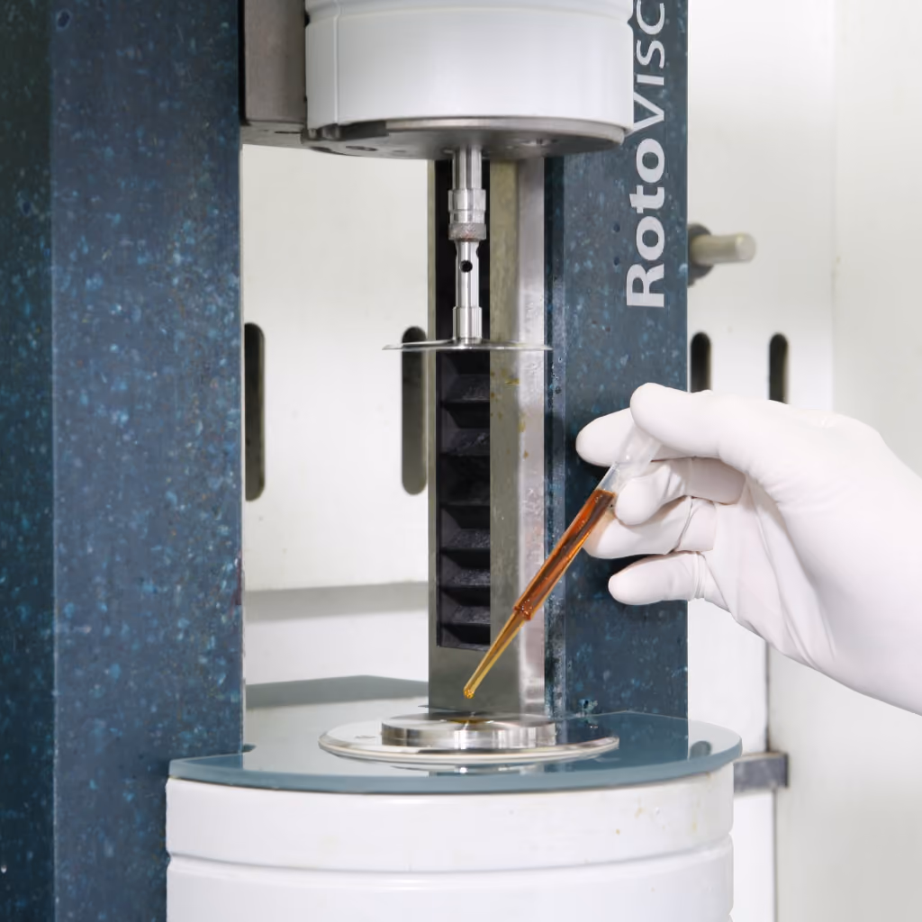 Hand in white glove holding dropper with brown liquid near a Roto Visco lab testing machine.