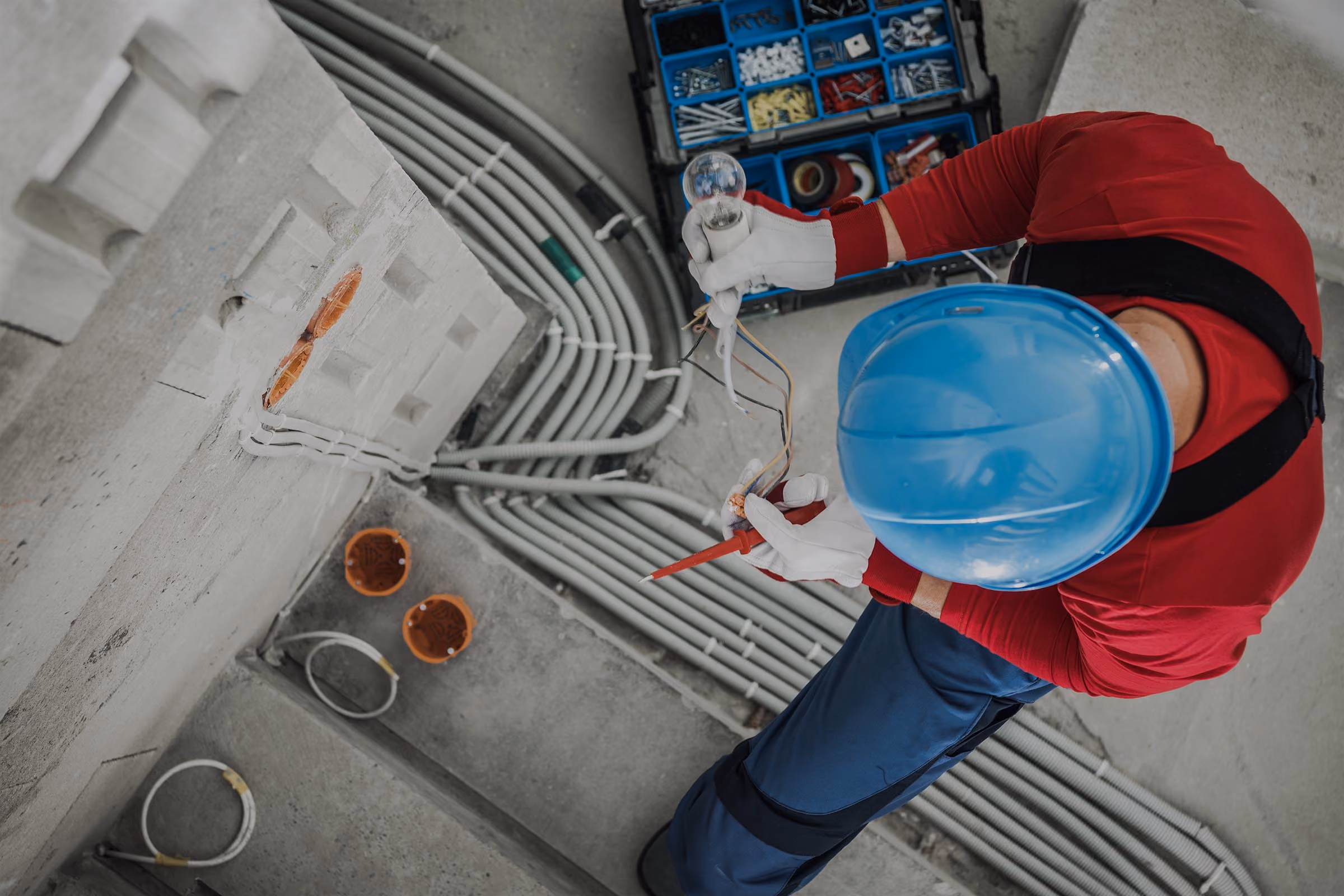 Electrician in blue helmet and gloves holding wires and a light bulb near electrical conduit pipes.