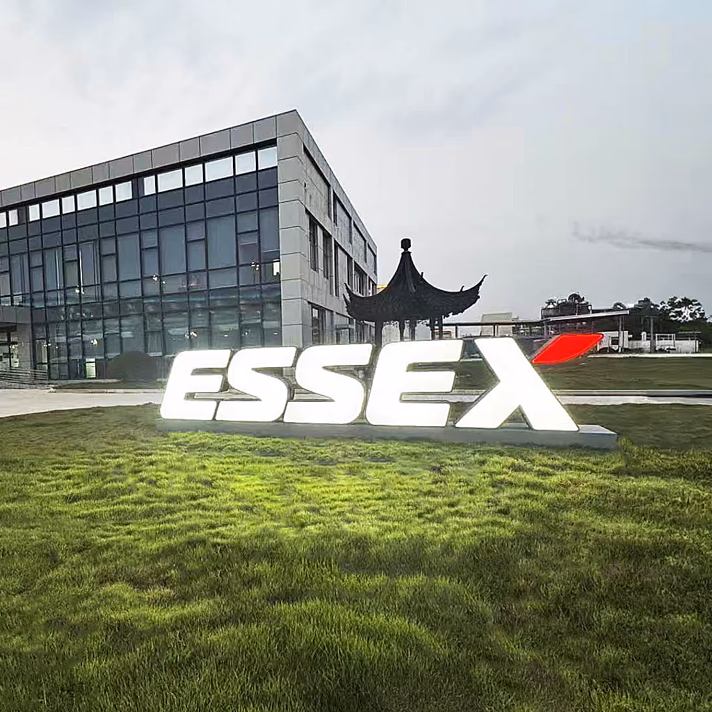 Illuminated large sign spelling 'ESSEX' with a red accent at the end, placed on green grass in front of a modern glass and concrete building.