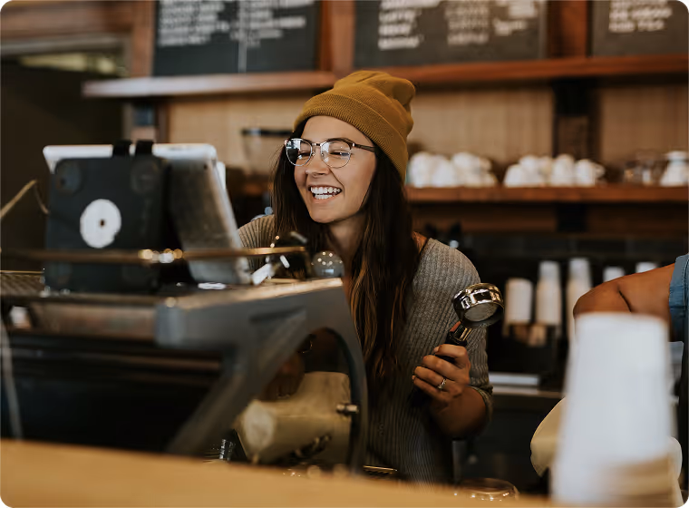 Smiling barista wearing glasses and a beanie holding coffee portafilter behind espresso machine in cafe.