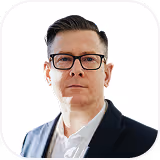 Man with dark hair and glasses wearing a dark blazer and white shirt on plain white background.
