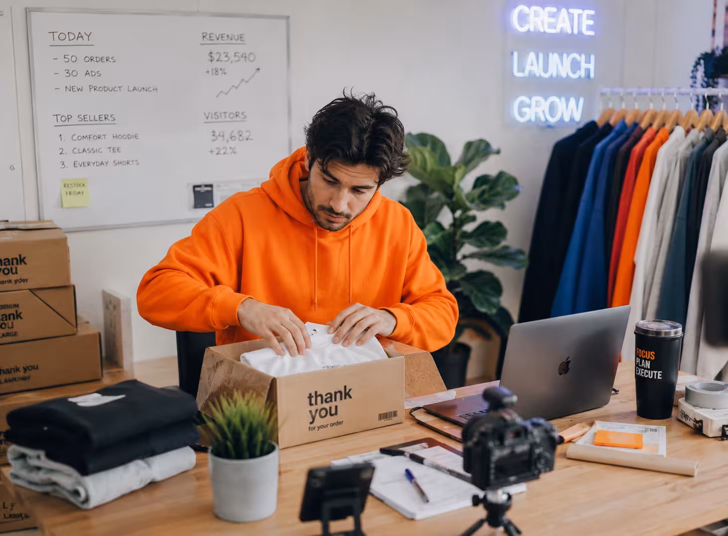 Man in orange hoodie packs clothing into a thank you box in a workspace with laptop and camera.