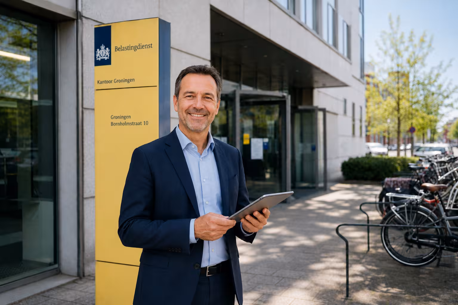 Smiling man in suit holding tablet outside office building with yellow Belastingdienst sign.