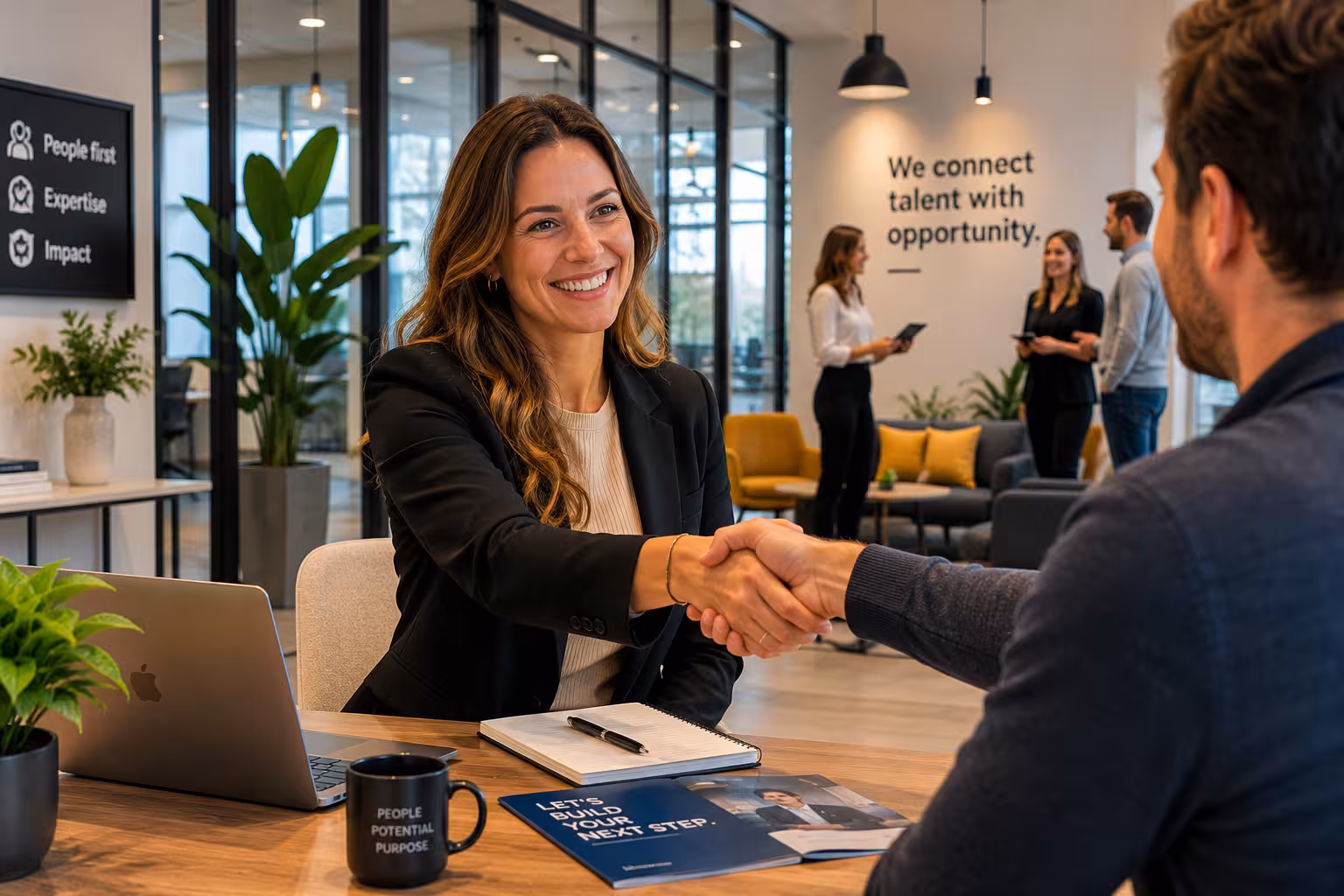 Smiling woman in office shaking hands with man, with people talking and recruitment quotes on walls.