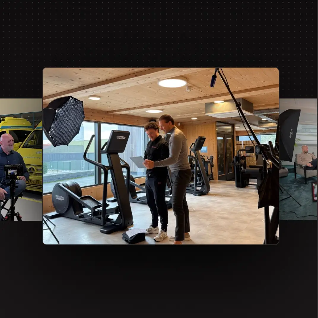 Two men in gym setting looking at a tablet with exercise equipment and filming lights around them.