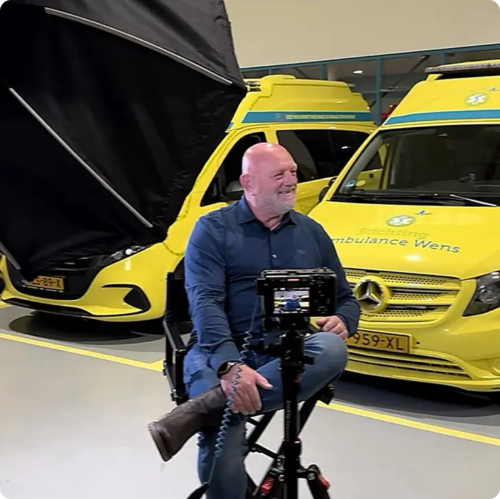 Smiling bald man with grey beard in blue shirt sitting on chair in front of cameras by two yellow ambulance vans.
