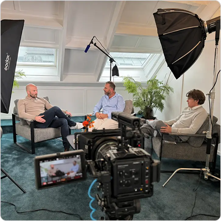 Three men seated indoors for a filmed interview with professional lighting and camera equipment.