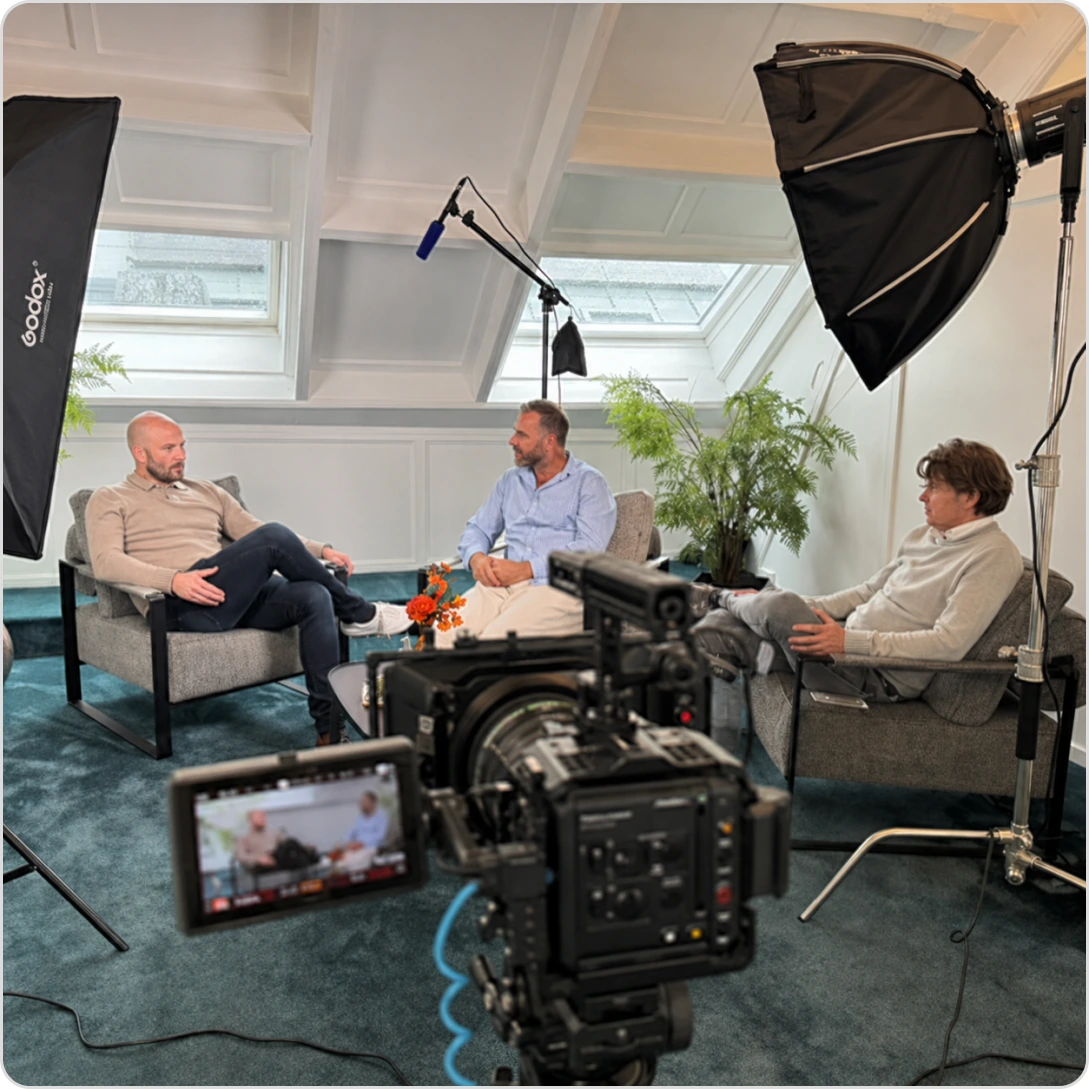 Three men sit in an indoor studio with camera and lighting setup for an interview or discussion.