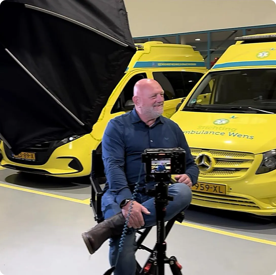Smiling man seated with legs crossed in front of a camera, with two yellow ambulances in the background.