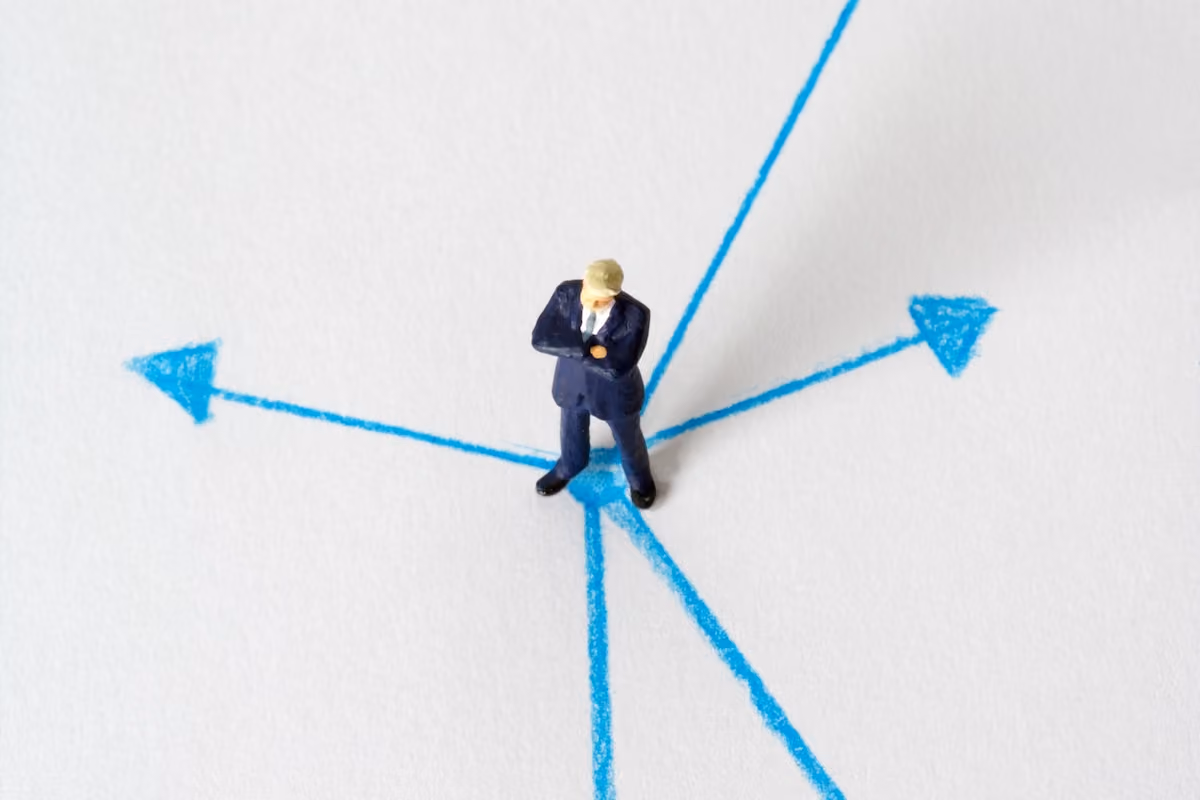 Miniature businessman stands at crossroads of blue arrows on white background, symbolizing choices.