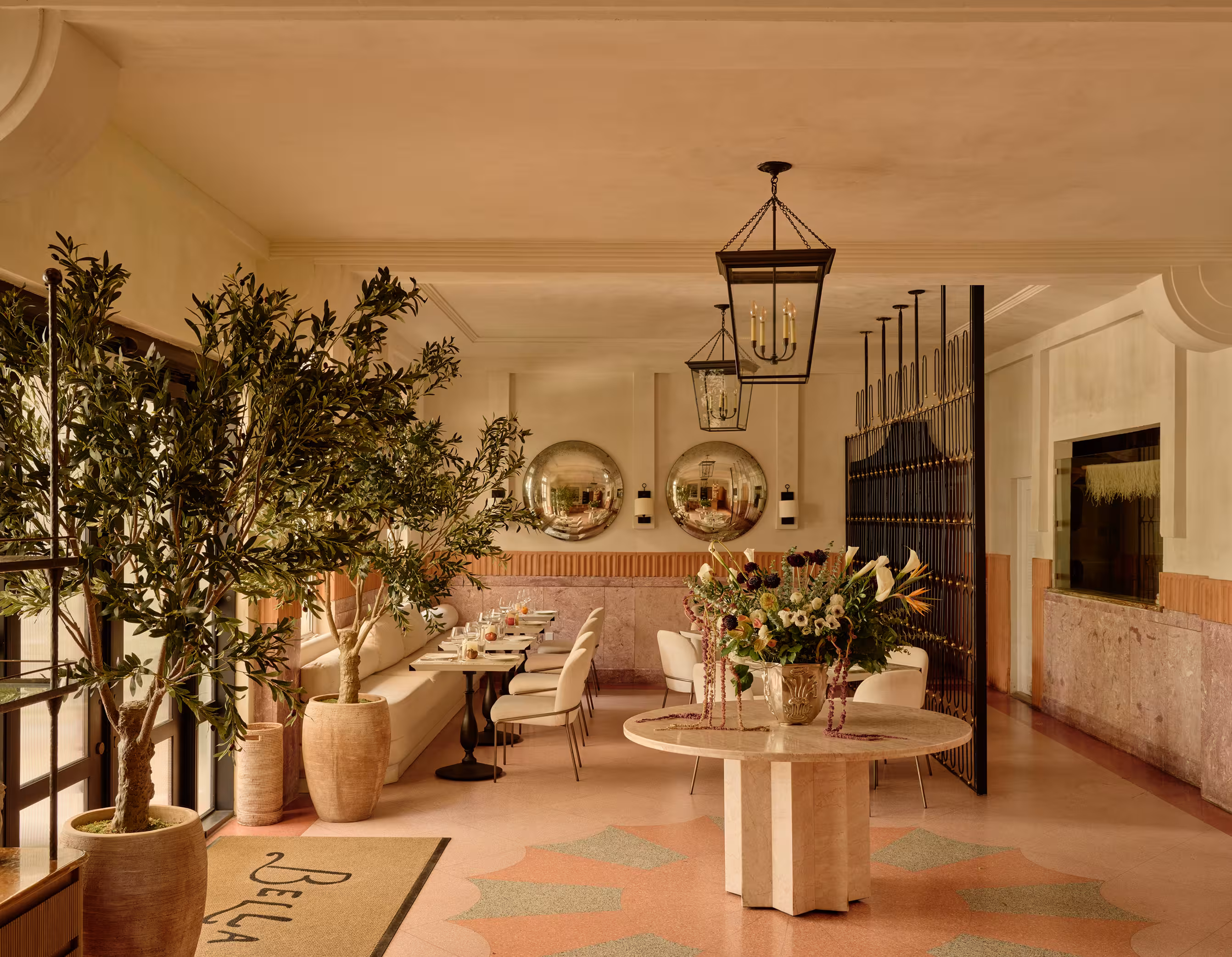 A home bar with glass wine shelving, wood stools, and an olive tree beside a fresh floral arrangement.