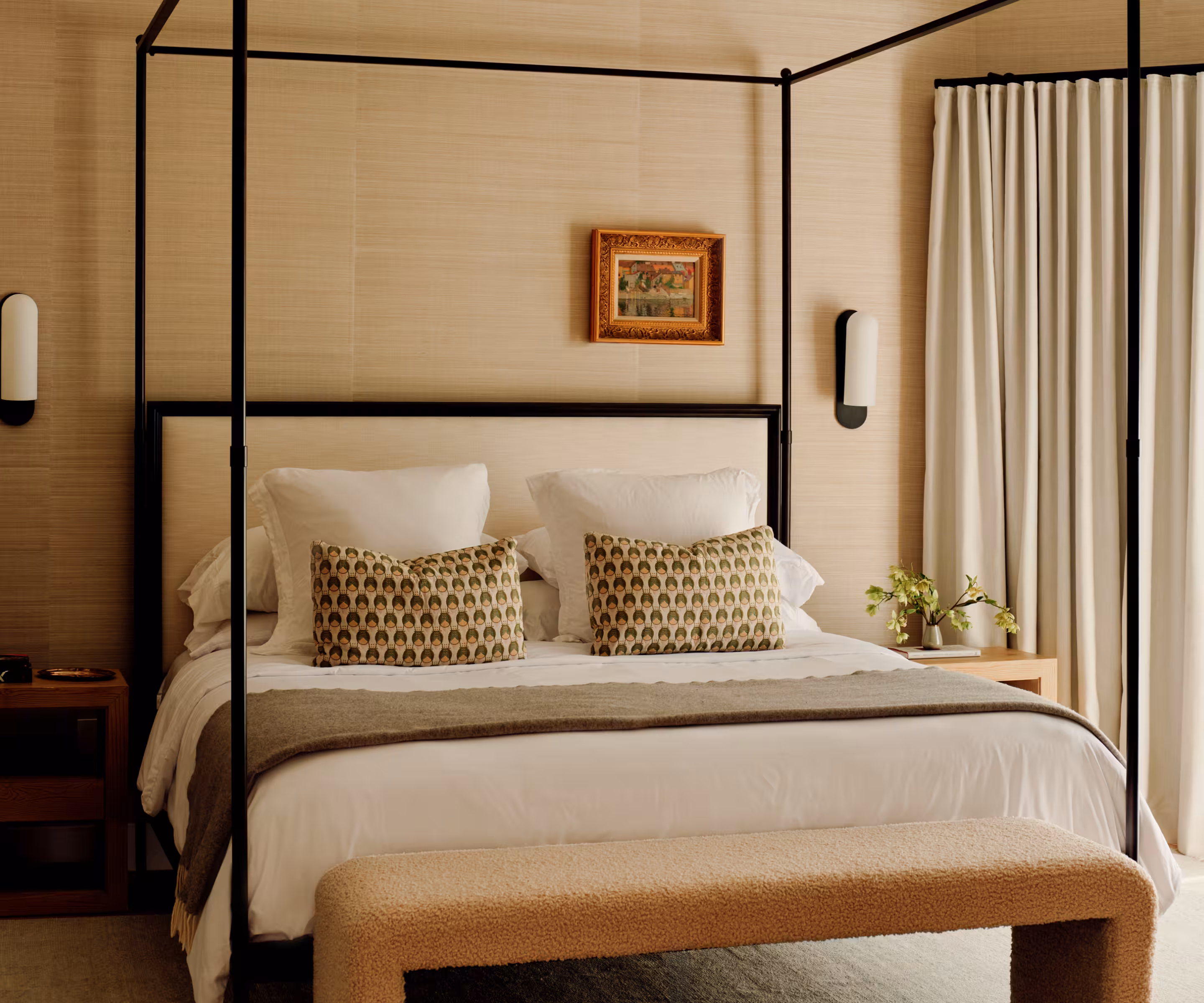 A four-poster bed dressed in patterned pillows against grasscloth-papered walls.